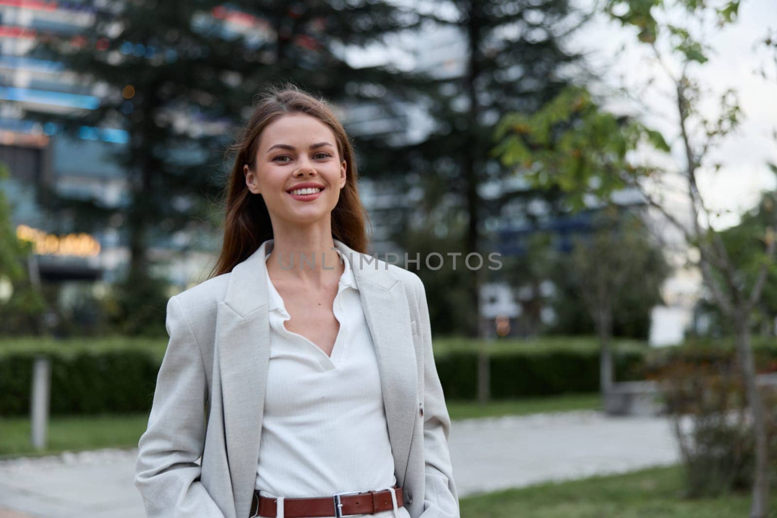 A confident woman in stylish business attire smiles warmly while standing in an urban park setting, reflecting professionalism and a positive attitude in her environment.