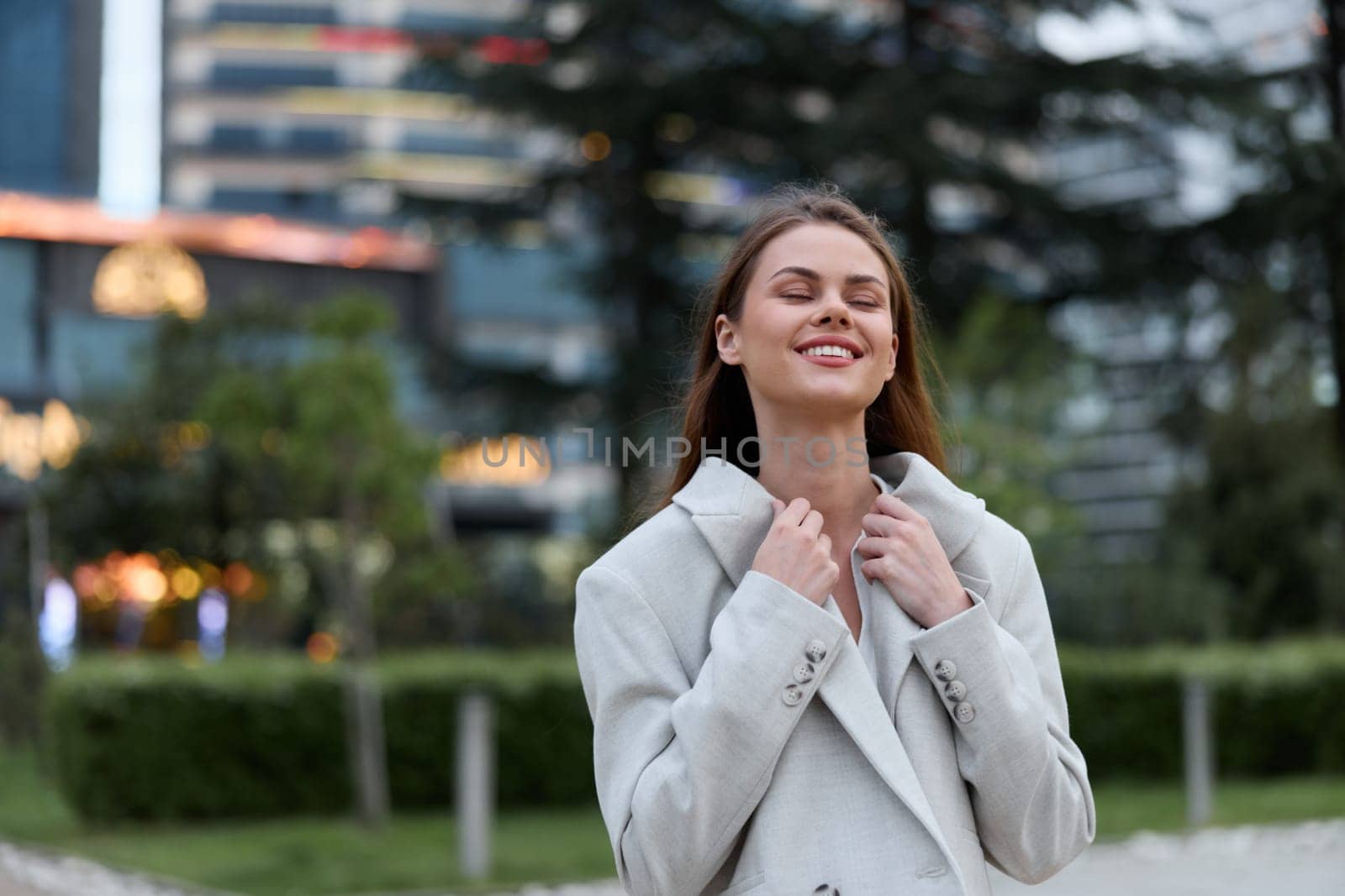 Young woman in light coat smiling and closing eyes with relaxed emotion outdoors in urban park setting, showcasing autumn style, modern lifestyle, and positive mood.