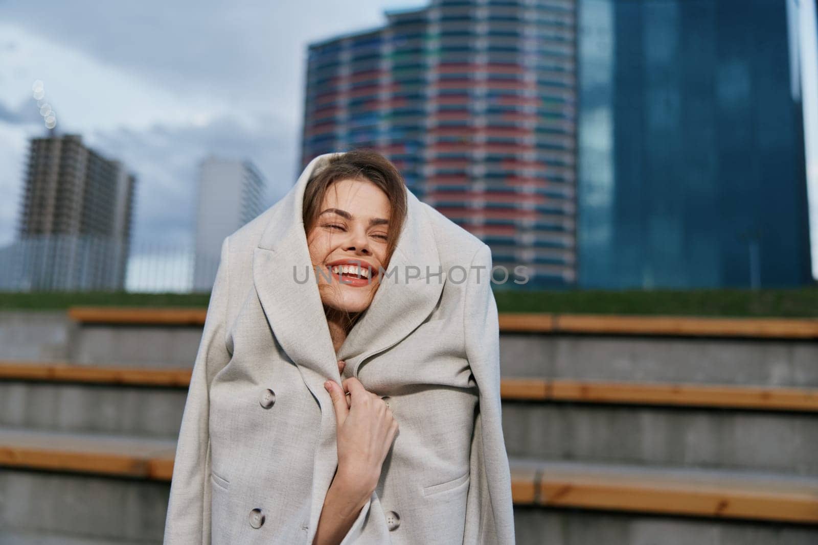 Happy woman wrapped in oversized coat smiles joyfully in urban environment during chilly autumn season, showcasing winter fashion and vibrant outdoor mood.