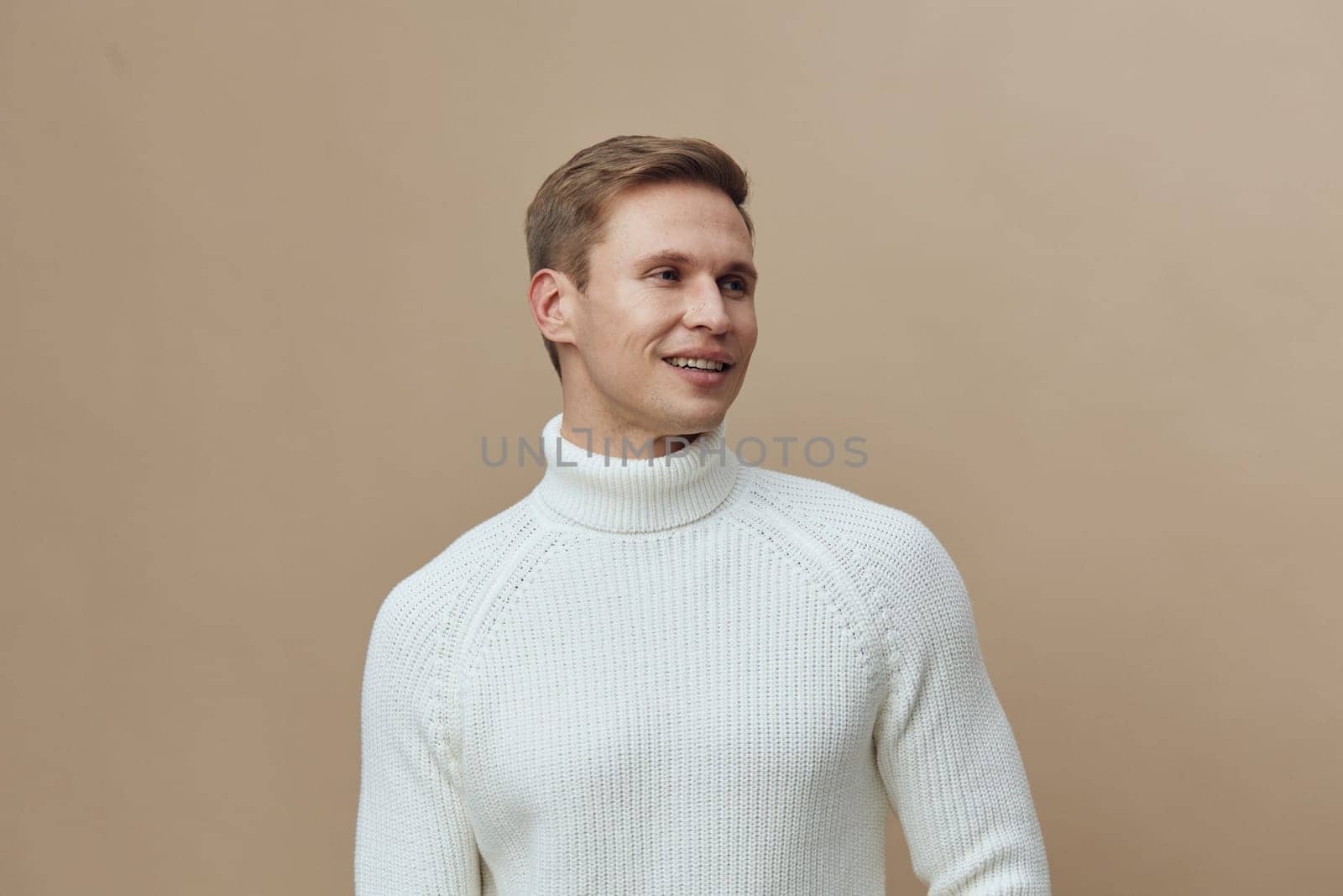 Smiling young caucasian man in white knitted turtleneck sweater looking to the side with positive emotion. Clean beige background emphasizes casual winter fashion and mental health gadgets concept. Healthy lifestyle and wellbeing.