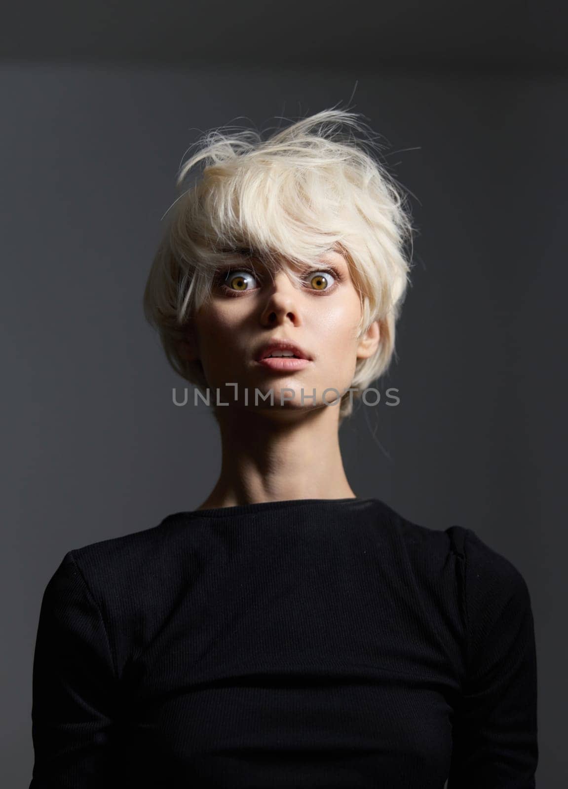 A young woman with striking short blonde hair and wide eyes, wearing a simple black shirt, captures an intense moment of surprise against a soft grey background.