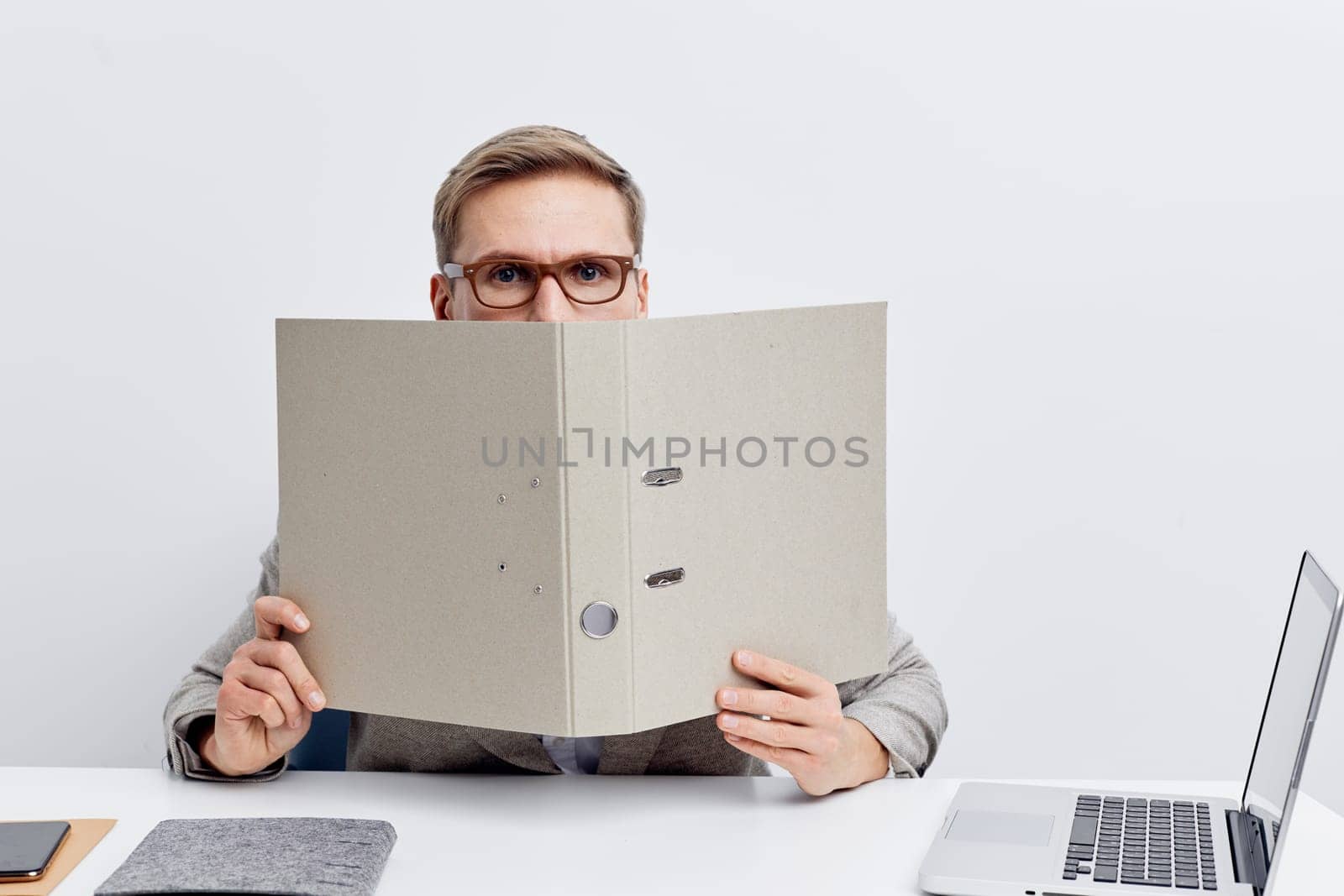 Young caucasian man working in cyberspace holding folder covering face while sitting at laptop wearing glasses and gray shirt isolated on white background. Technology, digital nomad, remote work, minimalistic studio background. Concept: digital nomad lifestyle