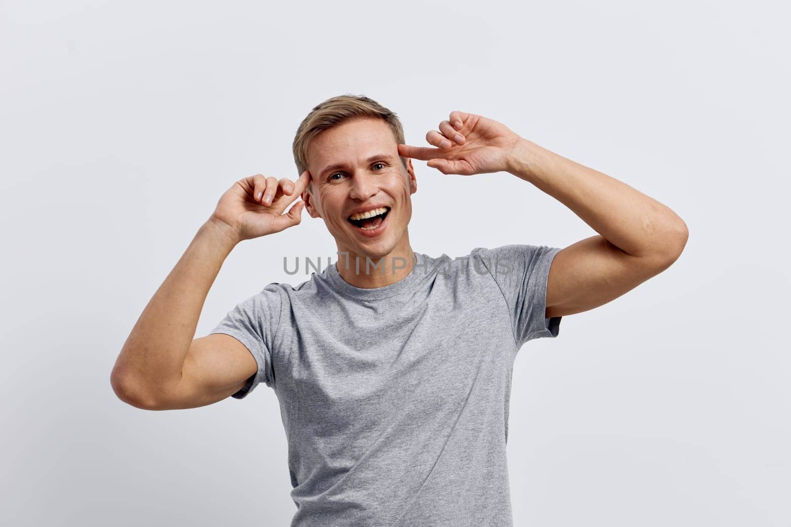 Happy young man with light brown hair in casual gray t-shirt smiling and playfully holding his ears, looking at camera against a plain white studio background. Positive emotion, joy, carefree mood