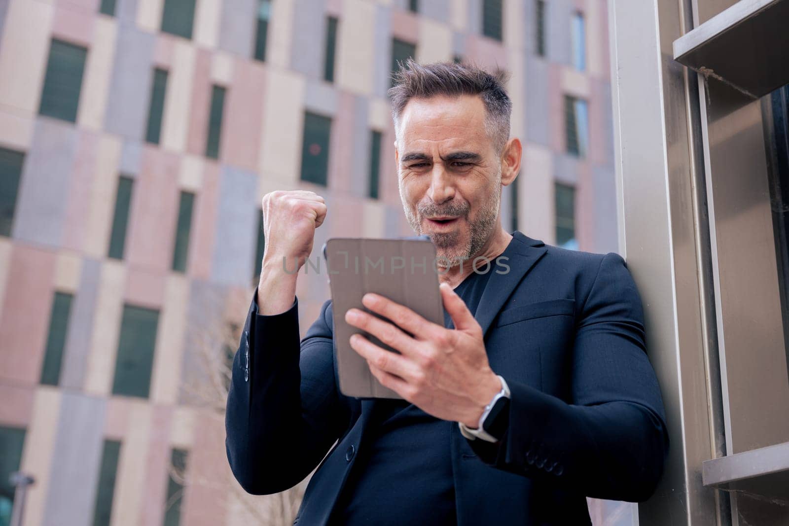 Confident businessman celebrates winning an online bet, holding a tablet and raising his fist. He smiles outdoors in a city setting. Concept of success, digital lifestyle, and modern finance.