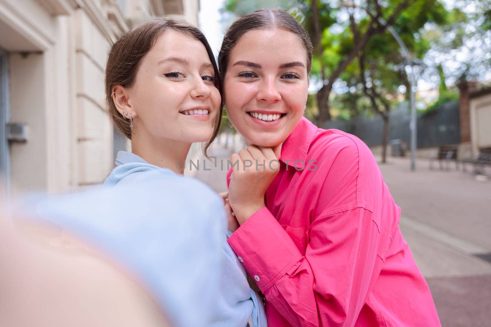Caucasian women smiling closely while taking a selfie together on a city street. Outdoor setting with trees and buildings, capturing a joyful and candid moment of friendship and connection
