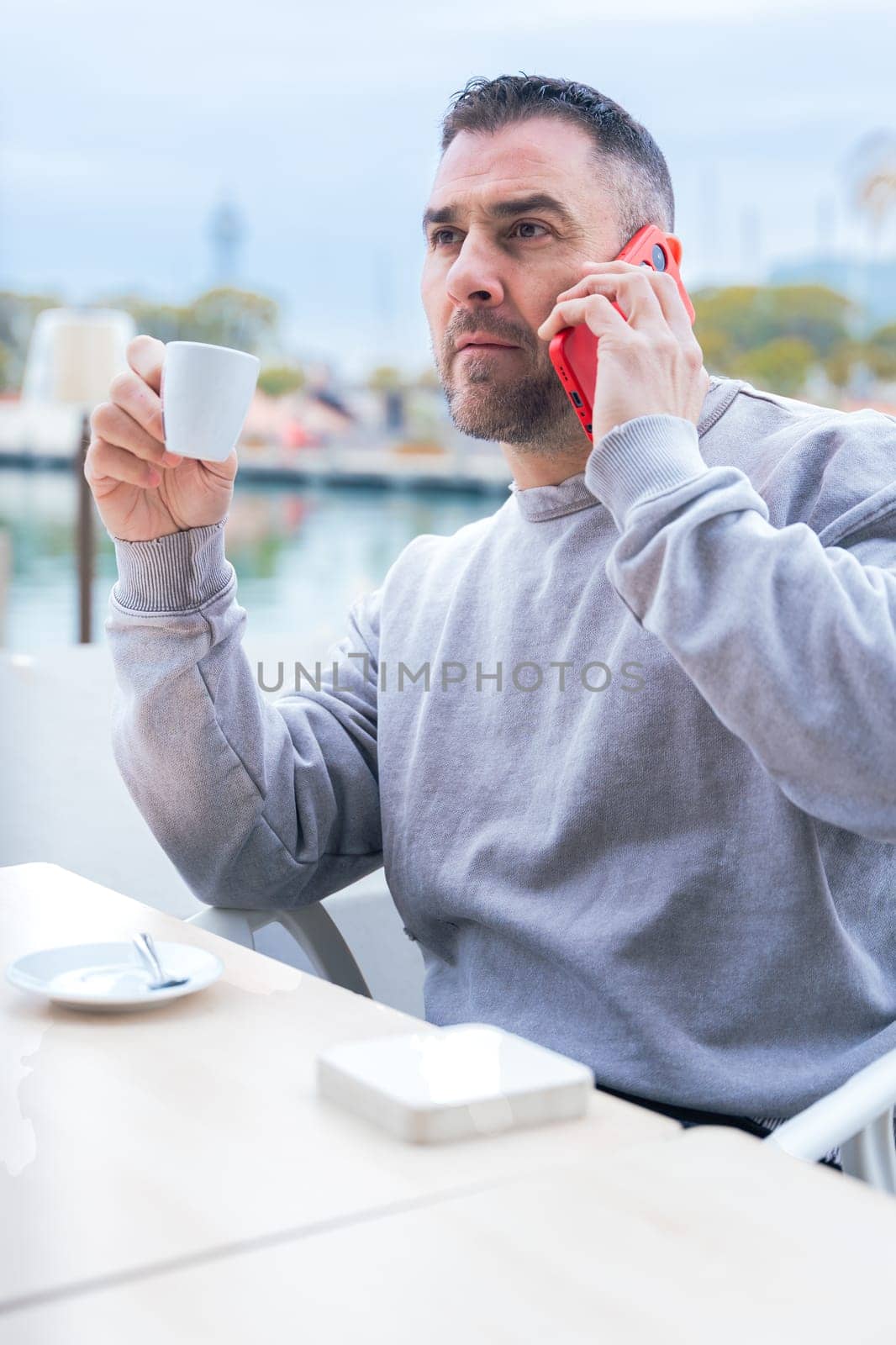 Man with short dark hair and beard drinks coffee while talking on his smartphone.He wears a casual gray sweatshirt and stands outdoors near the water,looking focused in a modern urban setting Vertical
