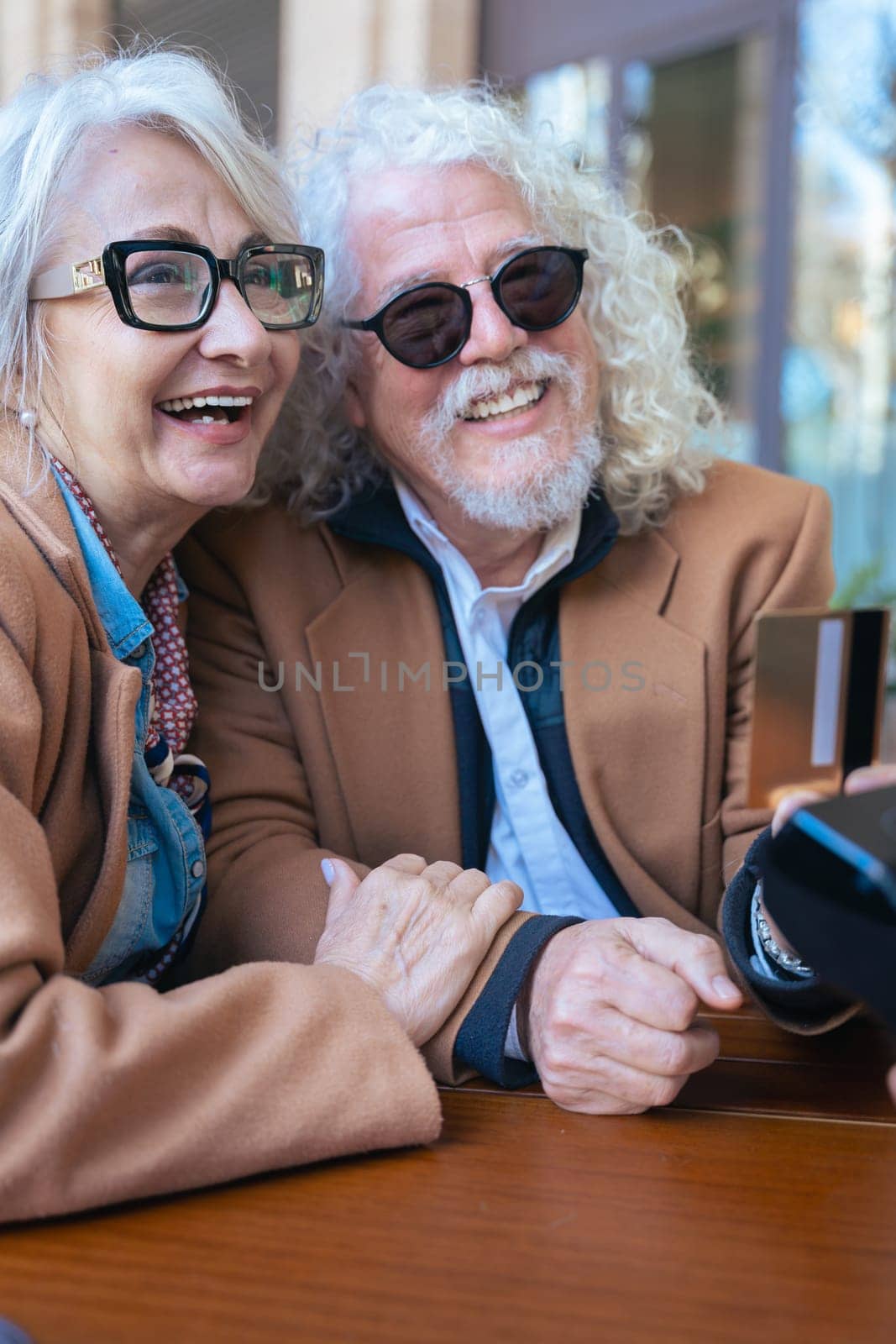 Joyful caucasian senior couple at an outdoor cafe making a card payment. Both wearing trendy outfits and sunglasses. Ideal for retirement, finance, lifestyle, and senior couple relationship concepts.
