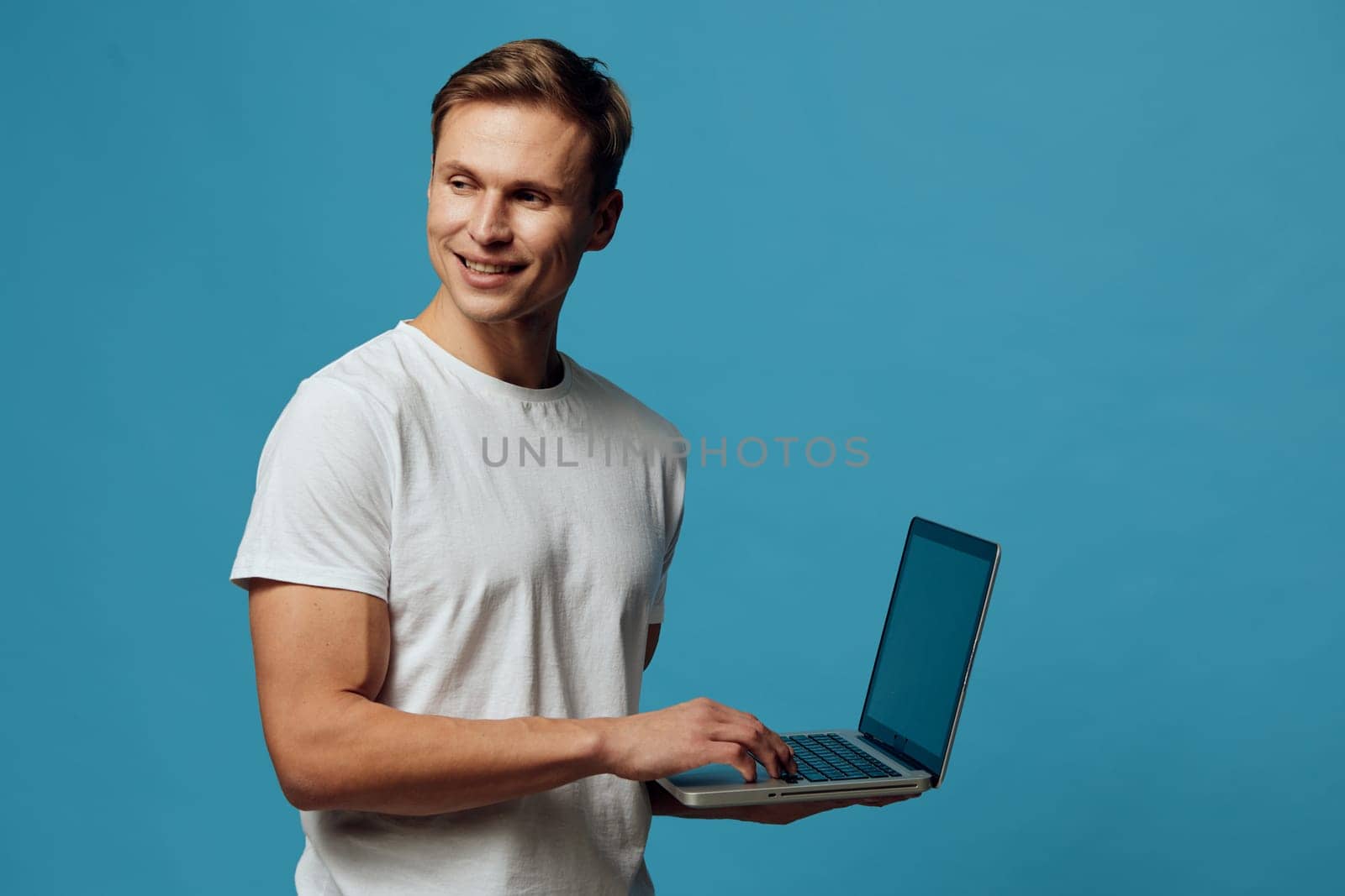 Happy man with short hair working on a laptop, looking at the screen and smiling, dressed casually in a white t-shirt, set against a plain turquoise background, studio shot emphasizing technology, communication, and contemporary lifestyle concepts