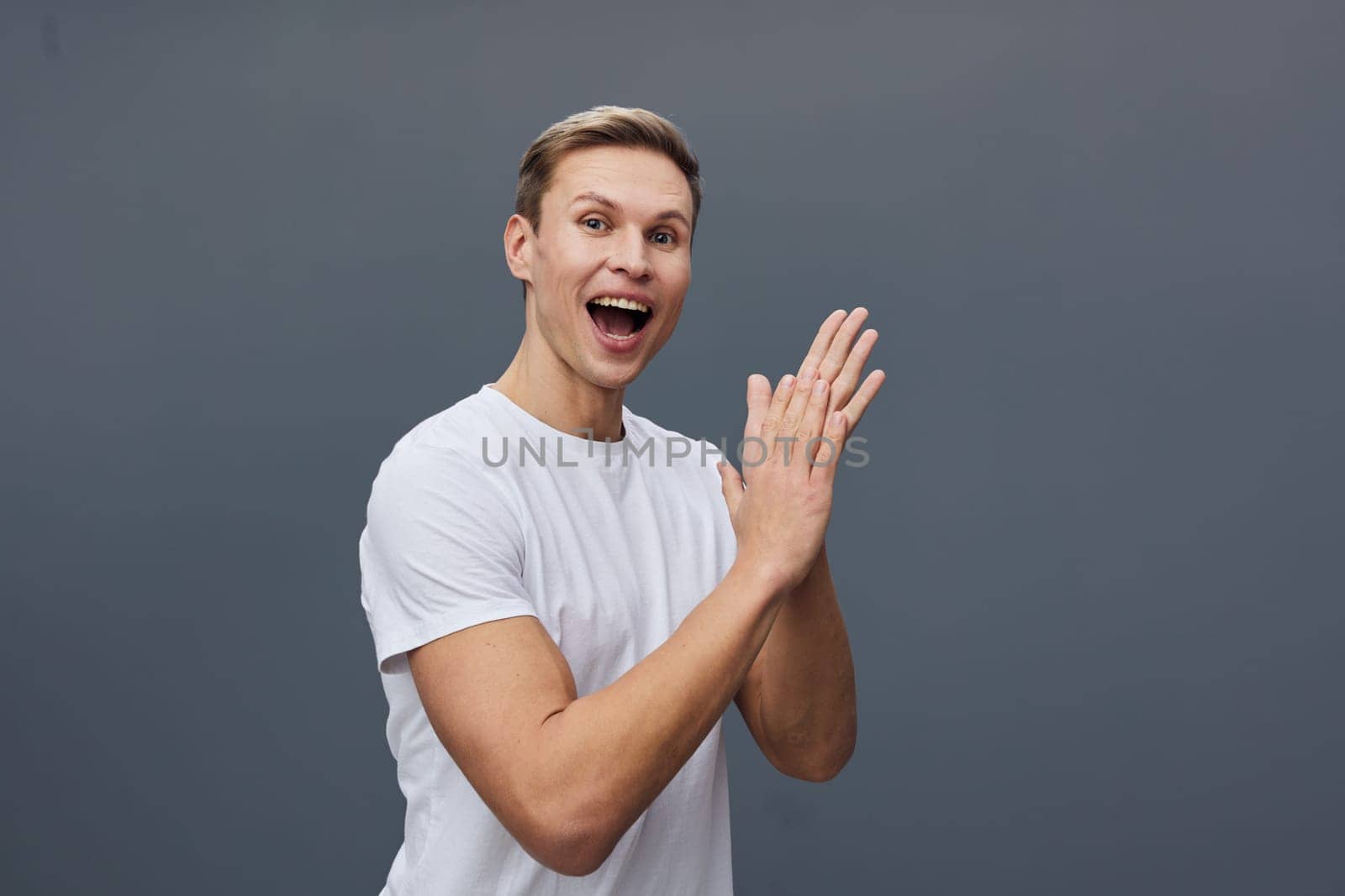 Happy cheerful man in casual white t-shirt showing excitement and joy, facing camera with hands together, on gray studio background. Concept: positive emotion and energy