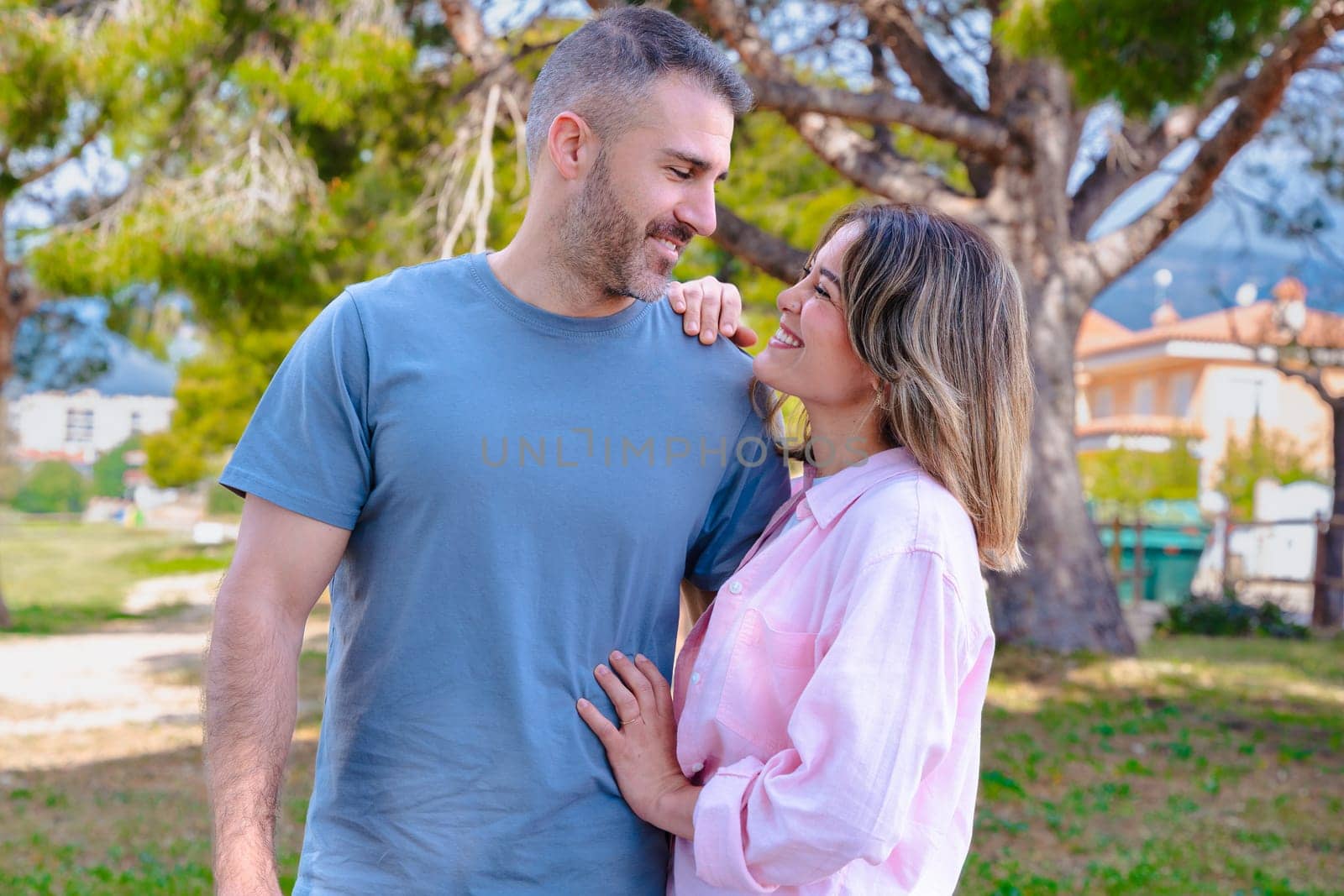 Young couple standing together outdoors, gazing into each other's eyes with love. Natural park environment, sunshine, green grass, and peaceful mood highlighting emotional connection and joy.