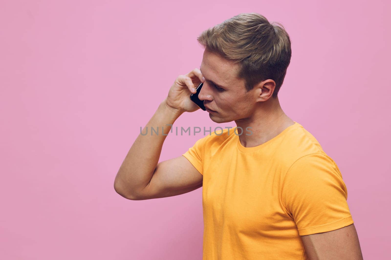 young man talking on smartphone wearing yellow t-shirt isolated on pink background close-up side view communication technology digital nomad lifestyle concept with mobile phone in hand