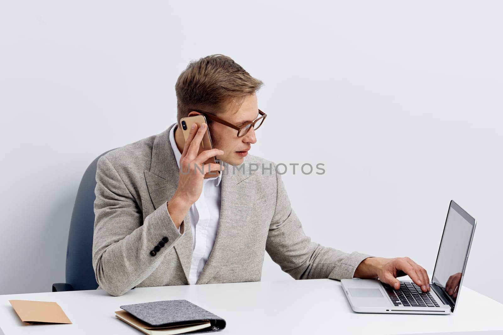 Middle-aged man dressed in business casual attire engaged in conversation on a smartphone while working on a laptop at a clean office desk, with a notepad and notebook, against a plain white background. Concept: professional communication, remote work.