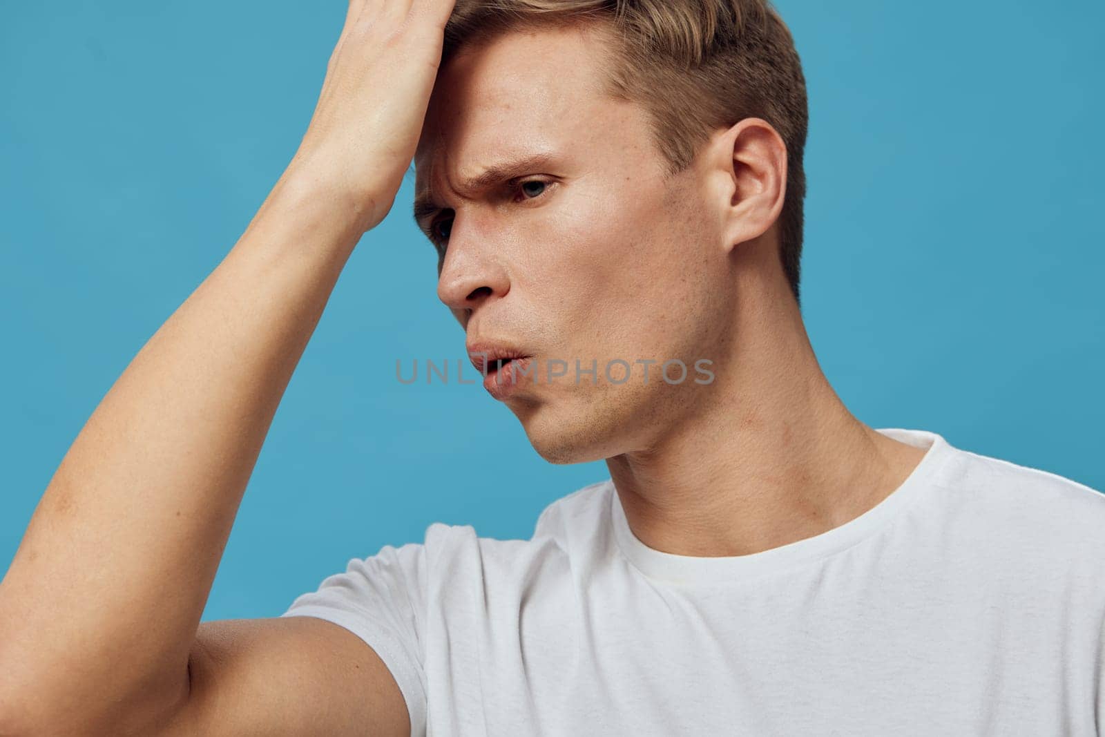 Confused or frustrated man with short brown hair, light skin, in casual white t-shirt, touching forehead with hand, deep in thought, expression of concern or stress, plain blue background, studio shot, emotional mood, human face.