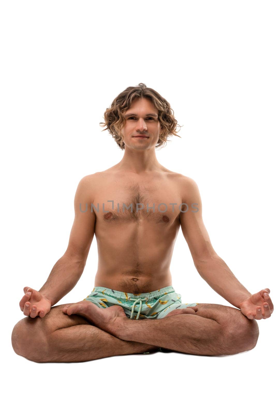 Full body barefoot young male in shorts with naked torso sitting in Padmasana pose and meditating during yoga session against white background