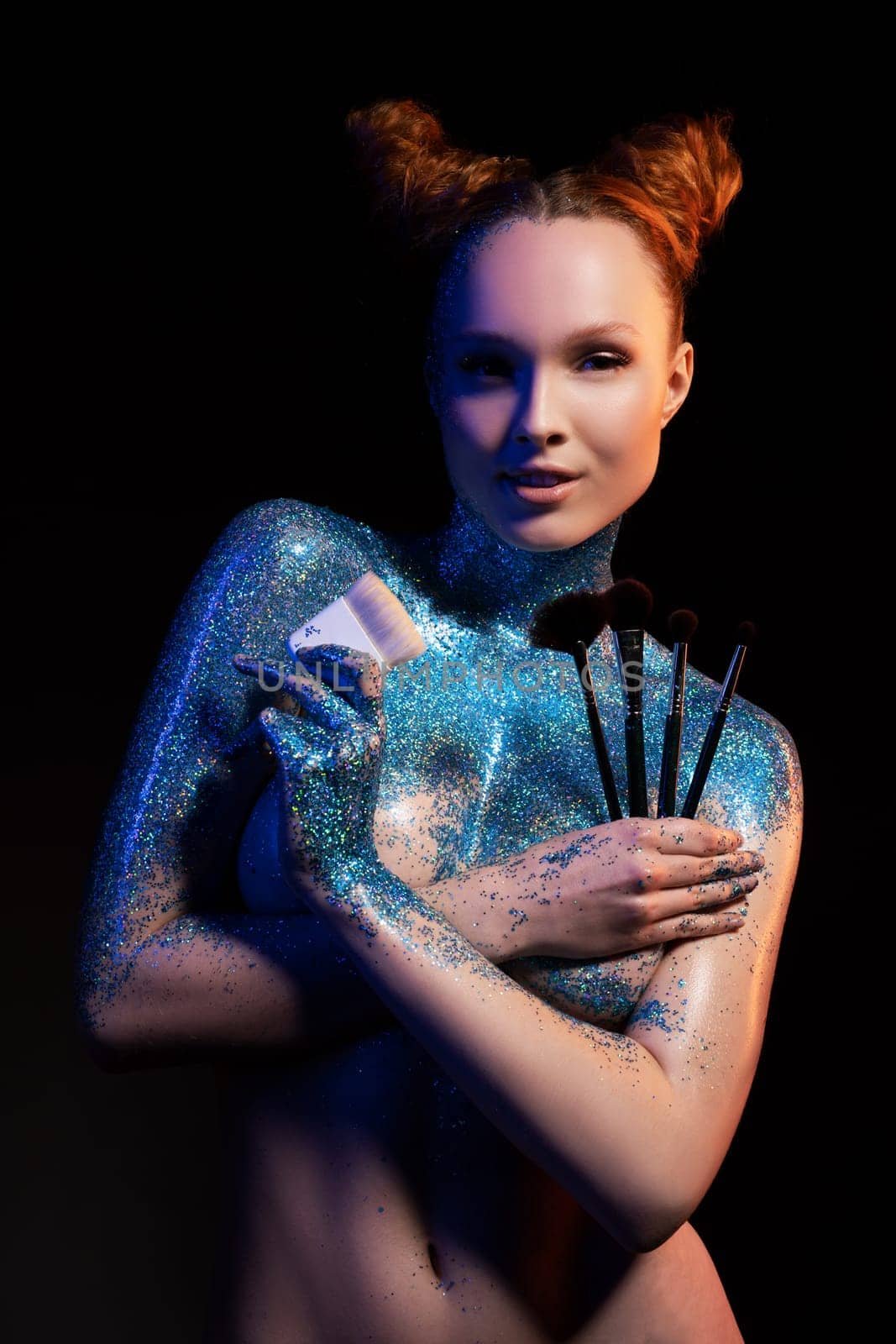 Self assured young female model with red hair topless painted with blue shiny glitter holding various makeup brushes and looking at camera