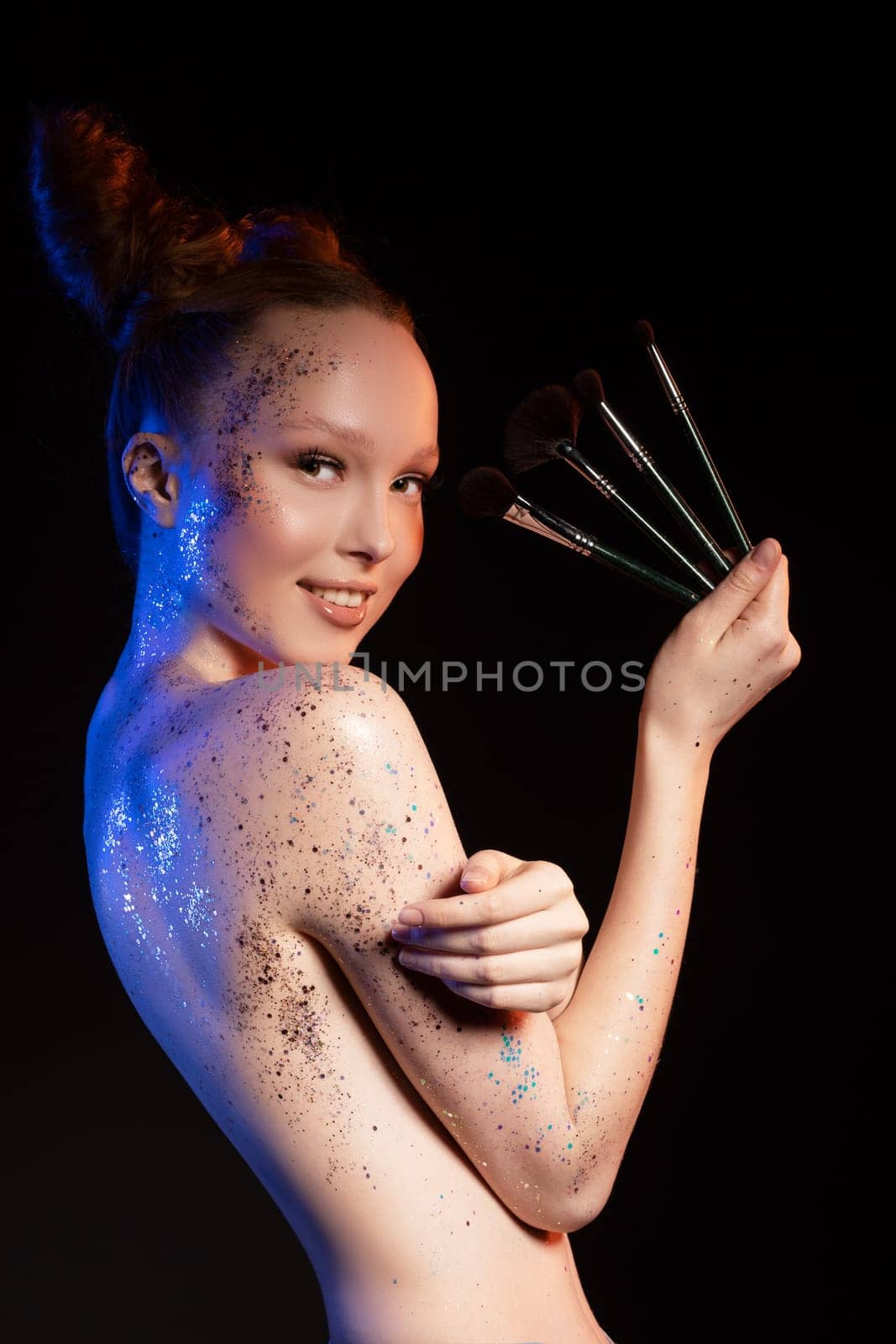 Sensual topless female with glitter painted body standing with makeup brushes in studio look at camera