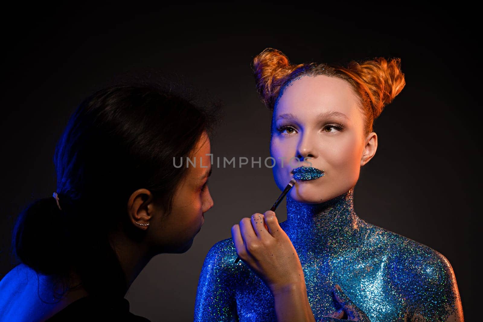 Side view of concentrated young female visagiste applying glitter on lips and naked body of confident model against black background in studio