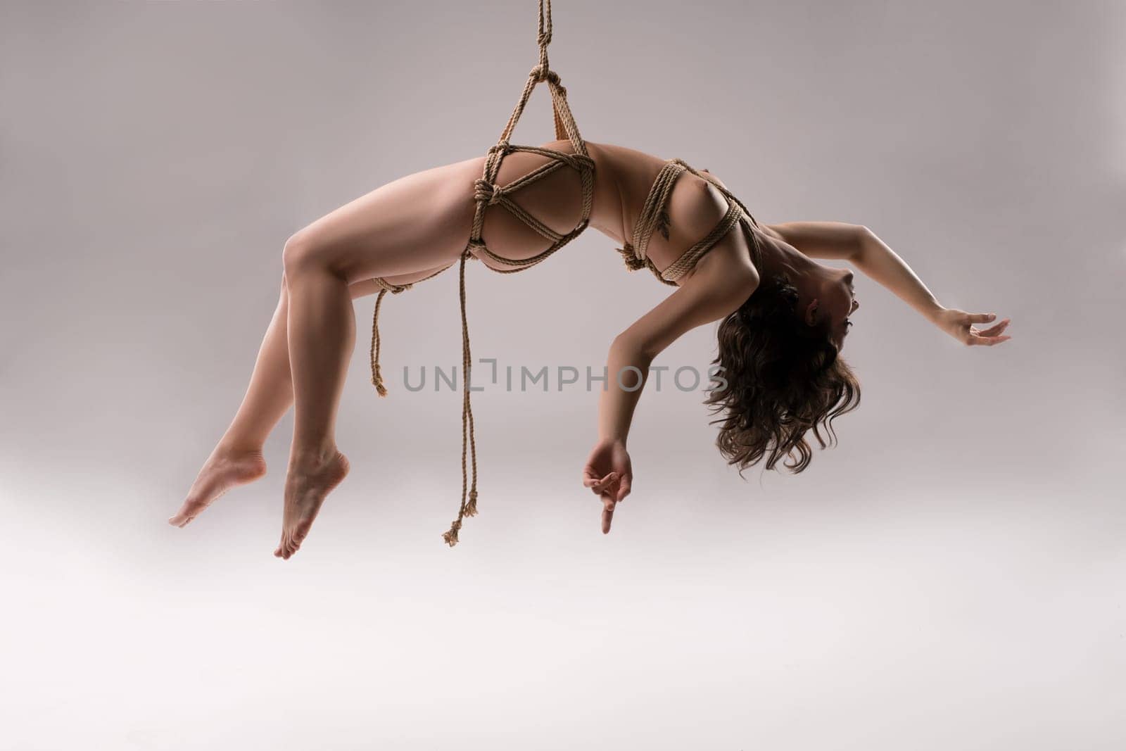 Full body of sensual naked female model tied up with rope against white background - bdsm shibari session