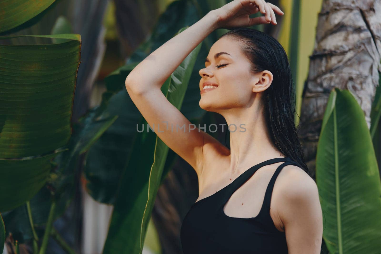 Woman in elegant black dress posing in front of palm tree with hands on head on tropical island resort