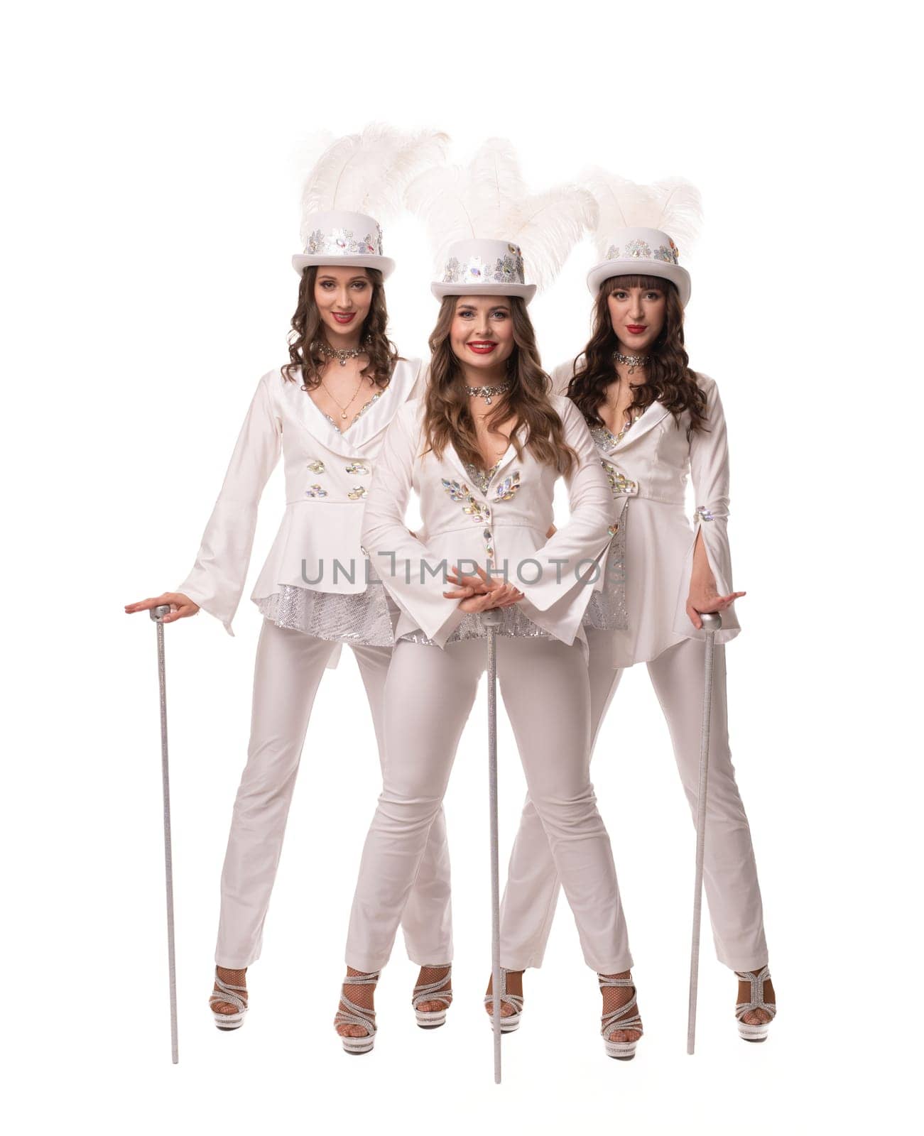 Full body of glad female in dance costumes and hats with feathers looking at camera with canes on white background