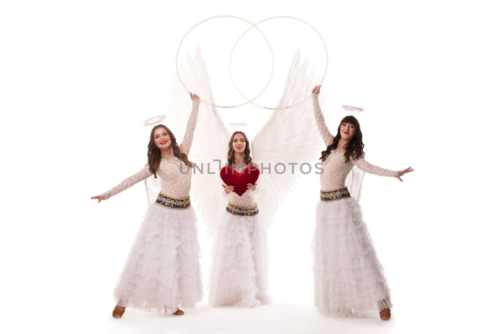Full body of female dancer in stylish angel outfits with wings and halos looking at camera with hoops and heart against white background