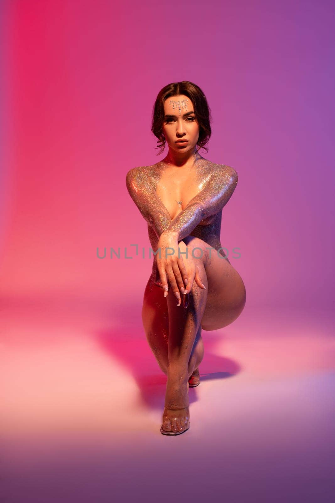 Full body of sexy young confident woman with dark hair and glossy paint on perfect body sitting on haunches and covering breast against pink background