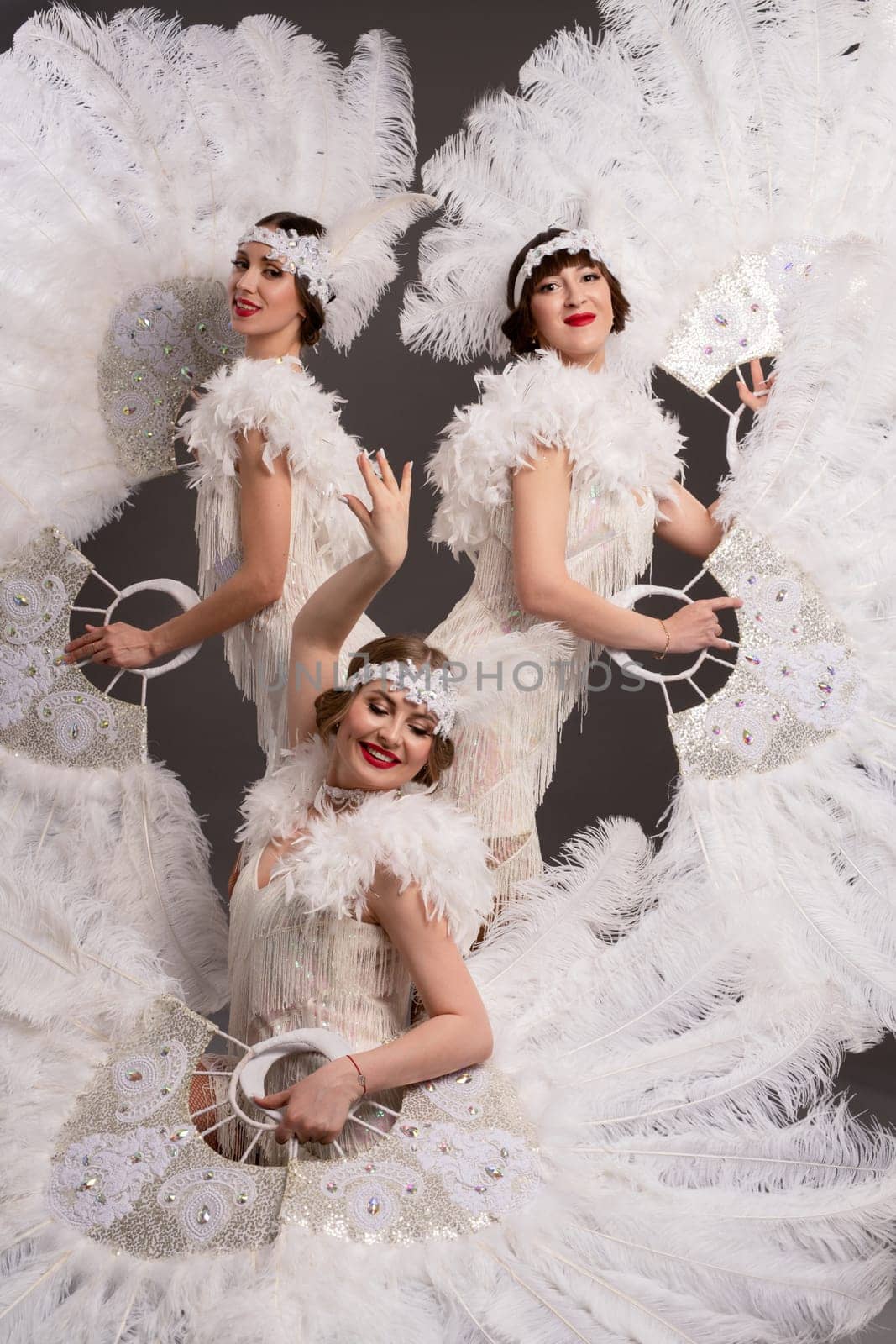 Beautiful female dancers in stylish burlesque costumes with headbands looking at camera with white feather fans in light modern studio