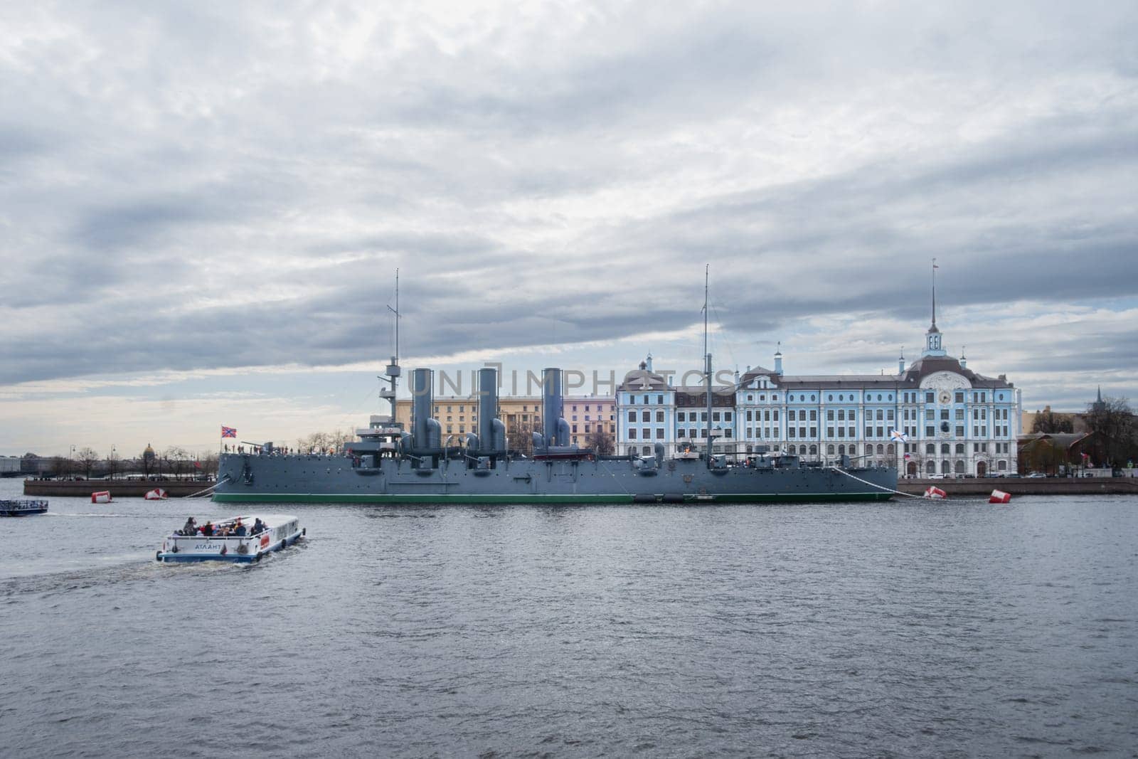Saint Petersburg, Russia - 02.05.25. Iconic cruiser Aurora in front of the Nakhimov Naval School on a cloudy day. A small boat passes by, adding a dynamic element to the scene.