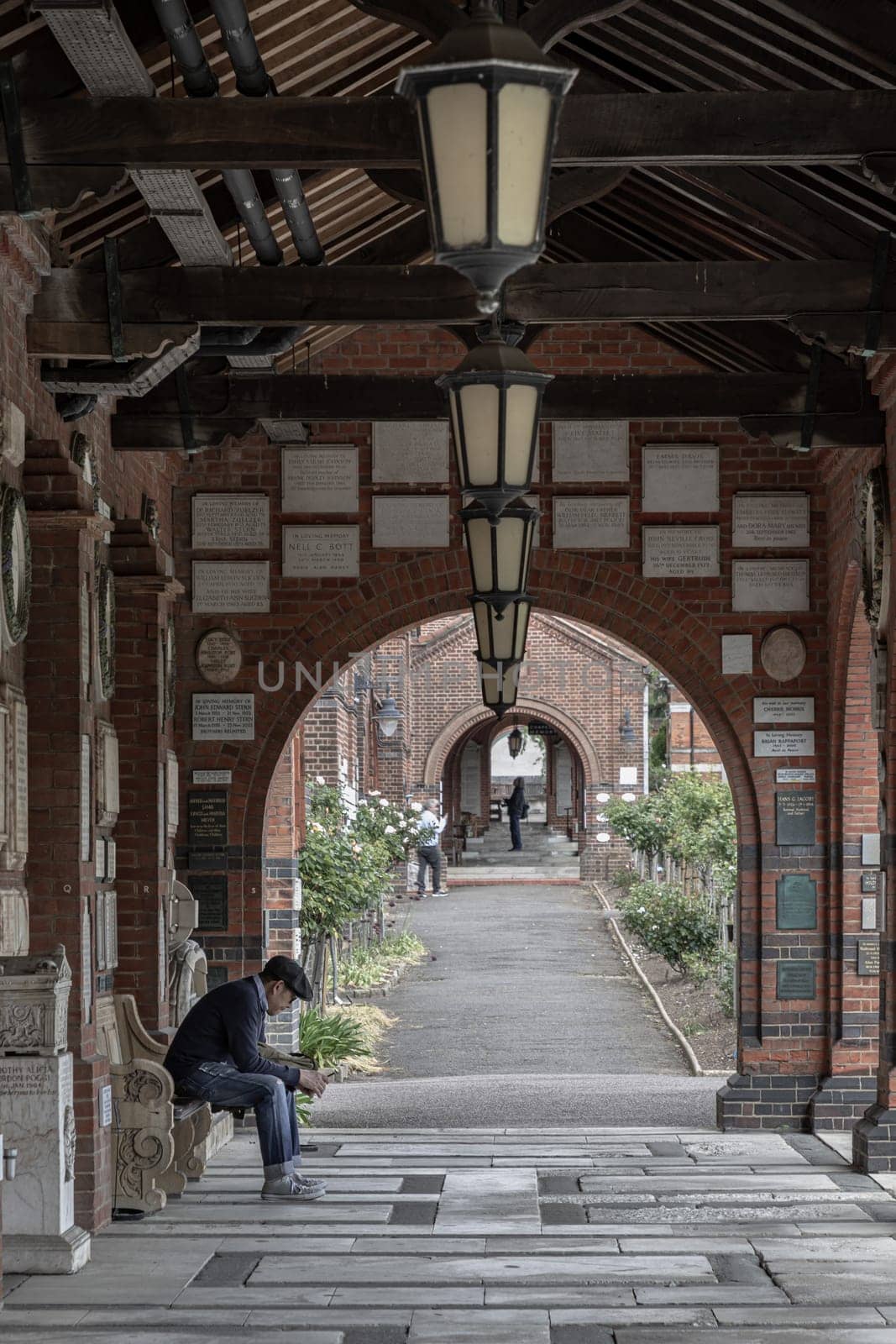 London, UK - May 08, 2025 - View of A man in long-sleeved shirt and jeans wear flat cap is sits on a wooden bench and looking down view messages at his smartphone under Hallway with Red brick columns and Antique lantern hanging from Wooden beams an Historic Building in Golders Green Crematorium and Mausoleum was the first crematorium to be opened at Hoop Lane. use it as your Wallpaper, Poster and Copy space, Selective focus.
