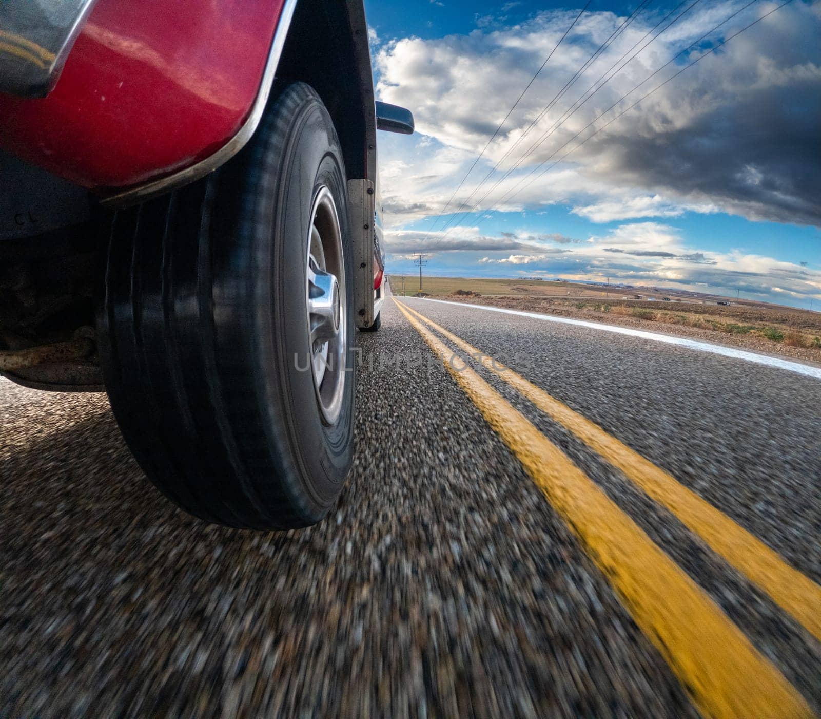 A red pickup truck is driving down a road with yellow lines. The road is empty and the sky is cloudy