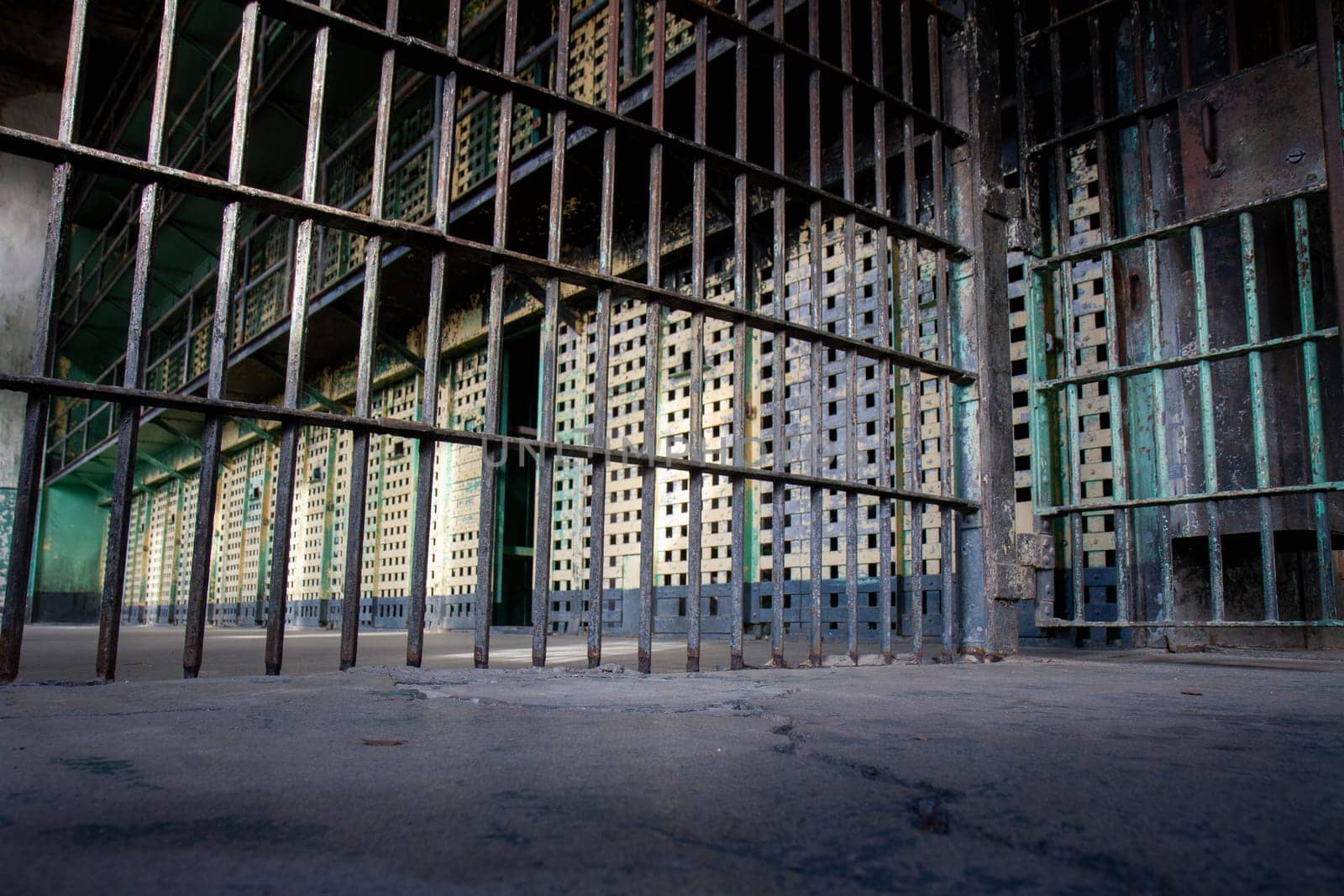 A prison cell with a metal fence. The bars are rusty and the cell is empty. Scene is bleak and somber