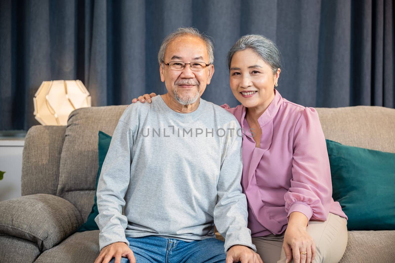 Portrait of Happy Asian senior couple greeting someone while sitting on a comfortable couch, old man and woman sit close together on a sofa in the house