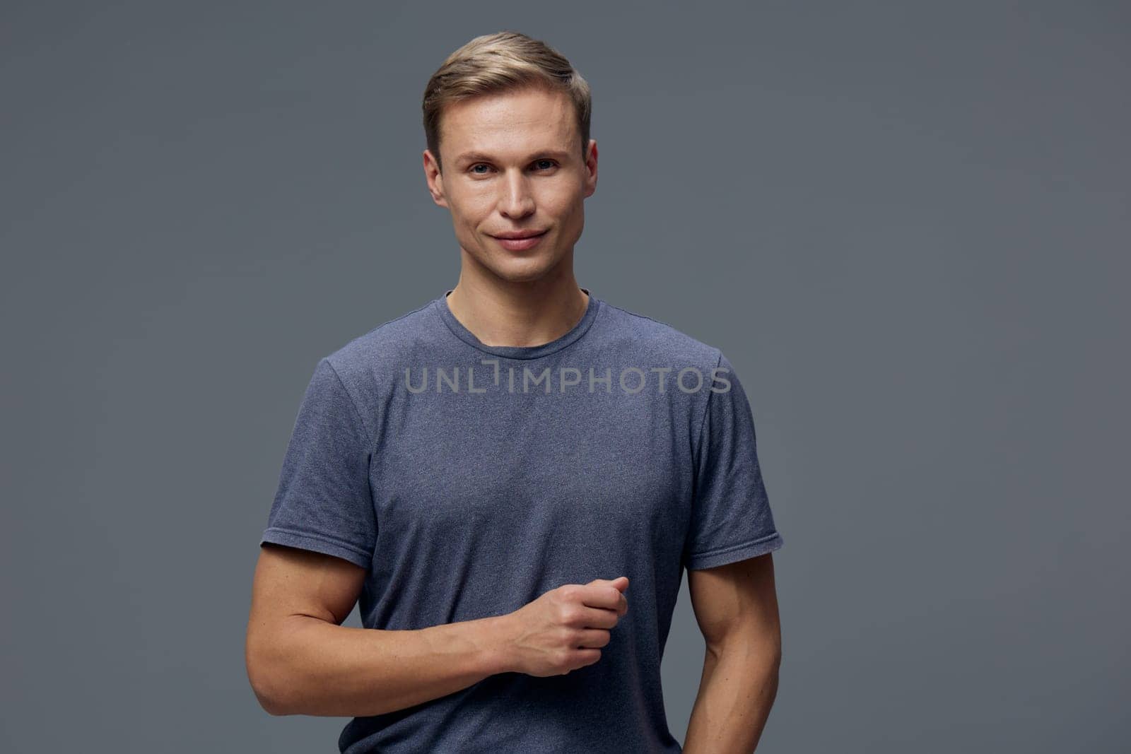 Friendly young man with light brown hair dressed in a simple blue t-shirt, standing confidently in front of a neutral gray backdrop. Calm, cheerful, positive mood, male model, casual attire, portrait photography, studio shot, lifestyle concept.