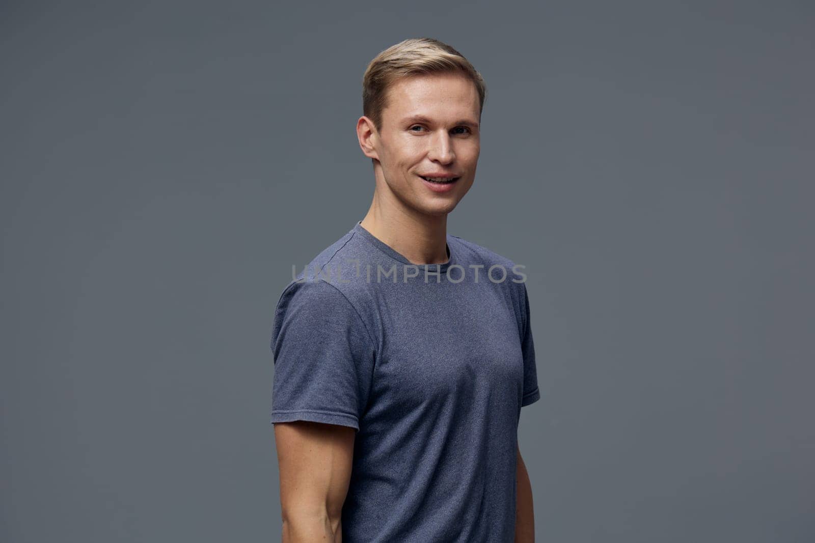 Happy young man with short blond hair dressed in a casual blue t-shirt, smiling confidently, posed against a neutral gray studio backdrop. Concept: positive attitude and relaxed style