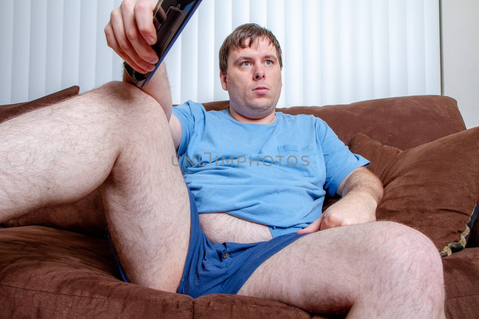A man is sitting on a couch with a remote in his hand. He is wearing a blue shirt and shorts