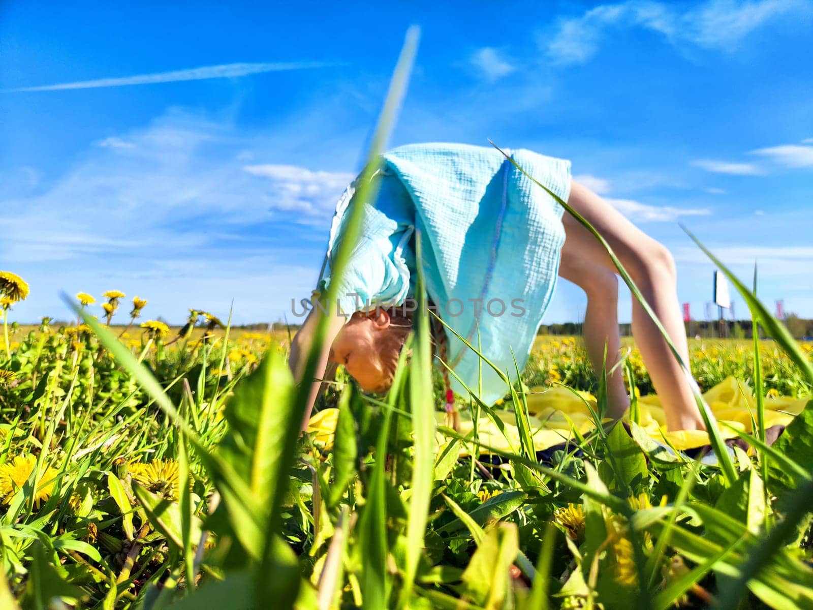 A young girl bends down to pick dandelions in a lush green field, surrounded by blooming flowers and clear skies.