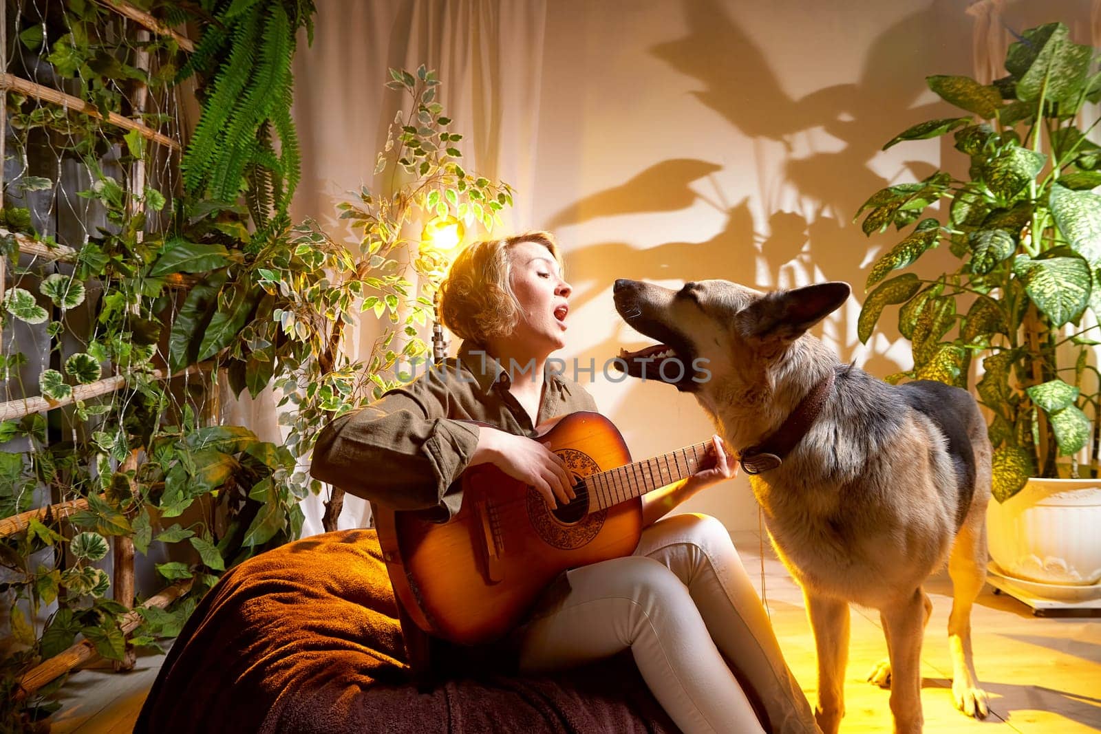 A woman sings joyfully to her dog while playing guitar in a warm, inviting room filled with plants and soft lighting.
