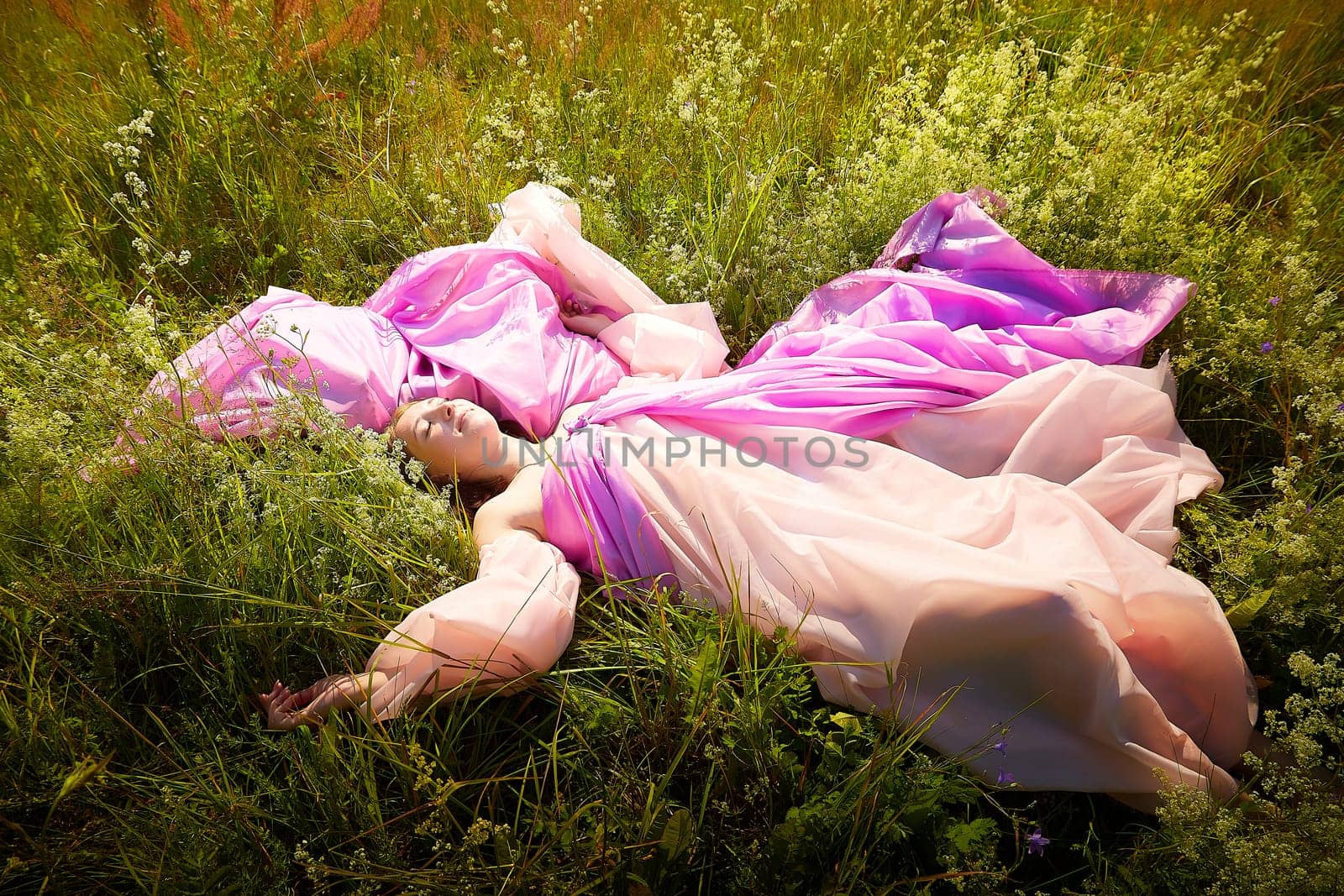 A woman lies on her back in a vibrant field, wearing a billowing pink gown that contrasts with the lush greenery and wildflowers around her.