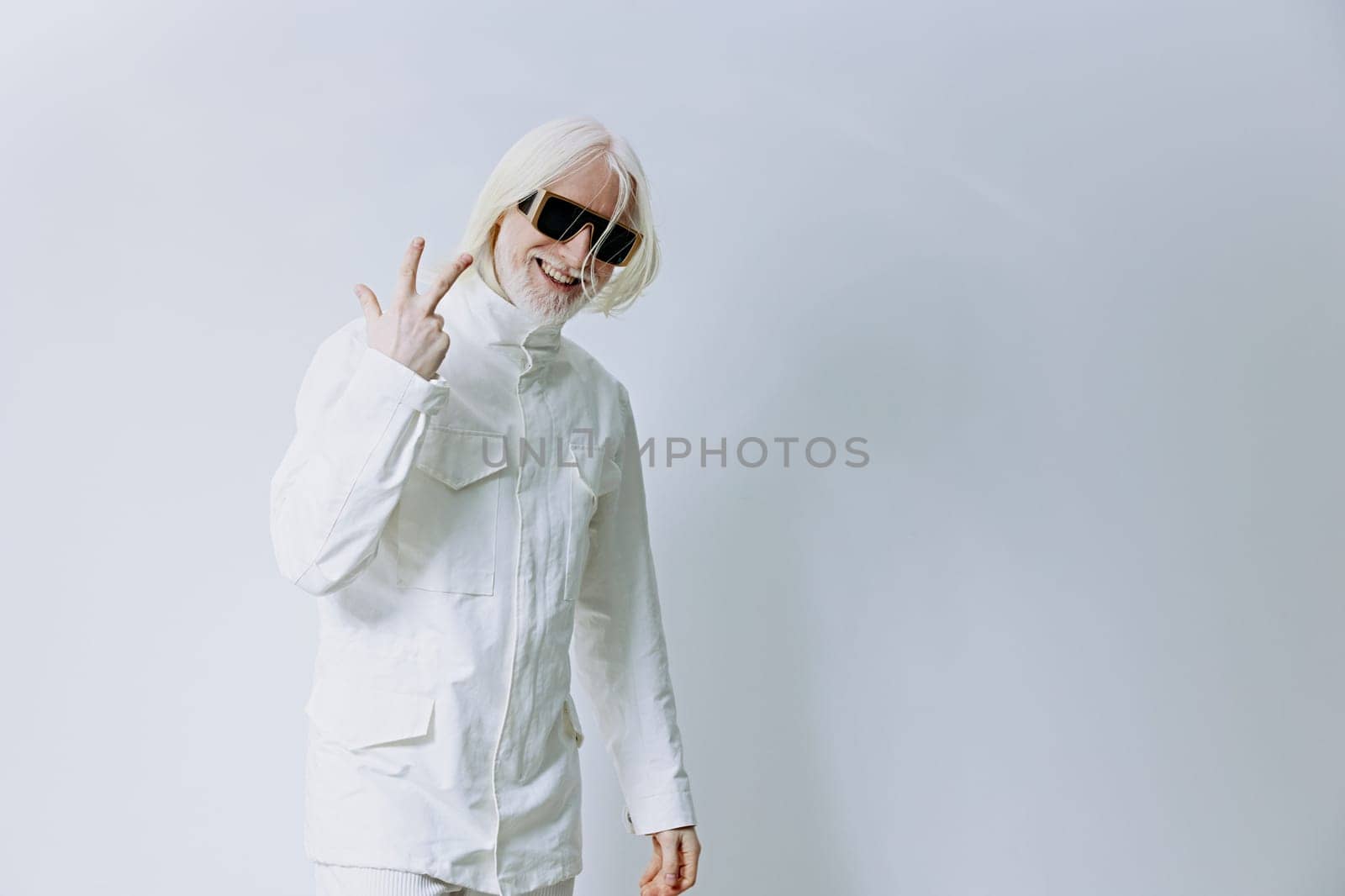 A trendy young individual dressed in an all white ensemble stands against a light backdrop, showcasing a peace sign with a smile, embodying a contemporary and positive lifestyle