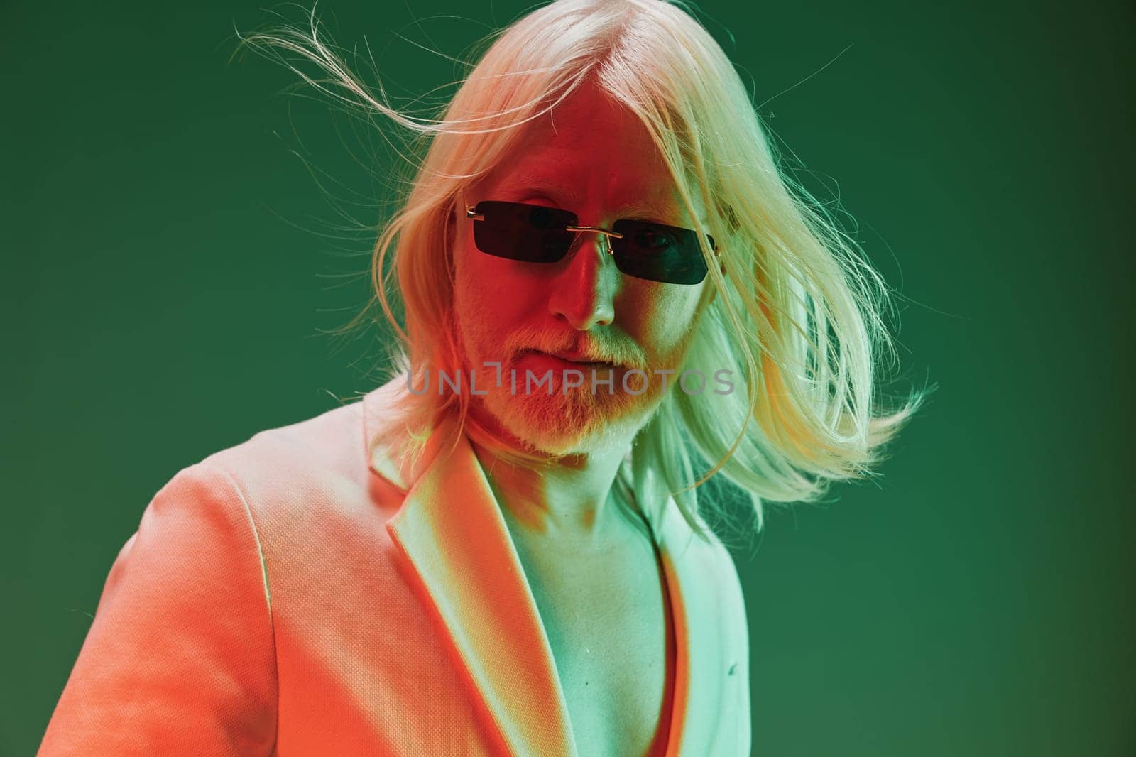 A striking image of a confident man with long blond hair and sunglasses, dressed in a sharp suit, set against a dynamic green and orange gradient background that enhances his stylish look