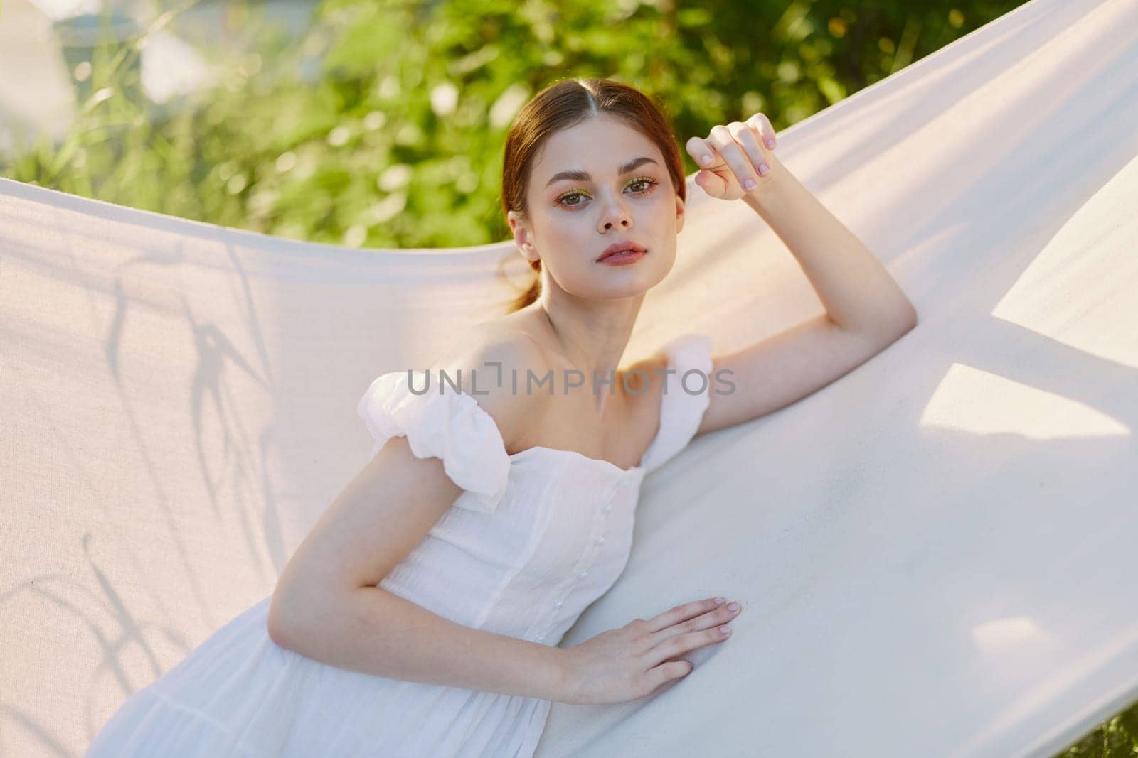 A serene young woman wearing a lightweight white dress reclines on a hammock, embodying relaxation and harmony with nature in a vibrant garden setting on a sunny day