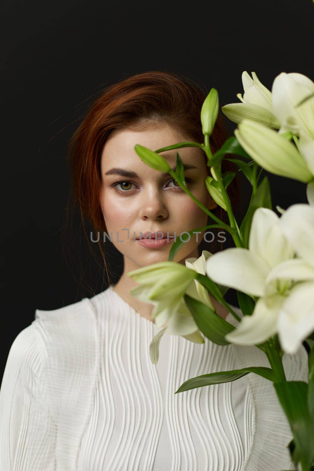 This portrait captures a young woman with reddish brown hair, dressed in a white blouse, holding white lilies The dark background enhances her natural beauty and elegance