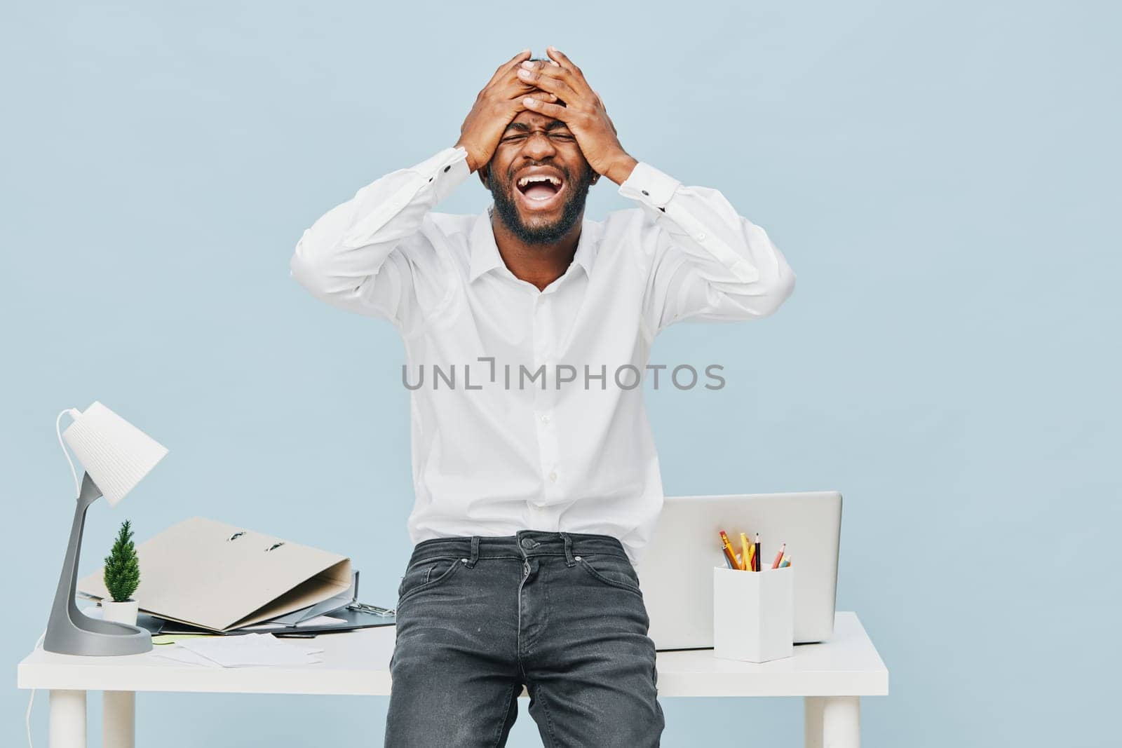 Happy man with dark skin and beard, wearing a white shirt and jeans, standing at office desk, covering face with hands, showing frustration or stress, in a clean workspace with pastel blue background. Concept: workplace emotion or work stress.