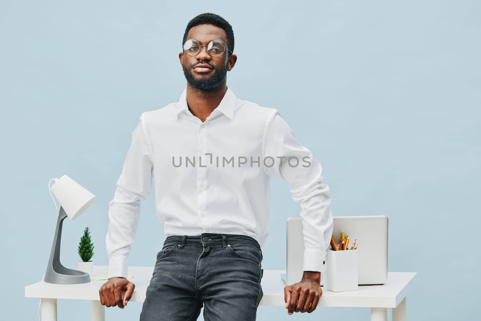 Stylish young man wearing a white shirt and black jeans stands confidently at a sleek desk with tech accessories and office supplies, set against a plain light blue background. Concept: professional workspace