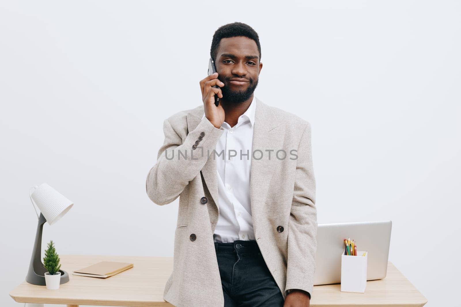 Confident man with beard using smartphone in modern office environment, wearing beige cardigan over white shirt, standing near desk with laptop, stationery and small plant, neutral plain background, corporate work scenario.