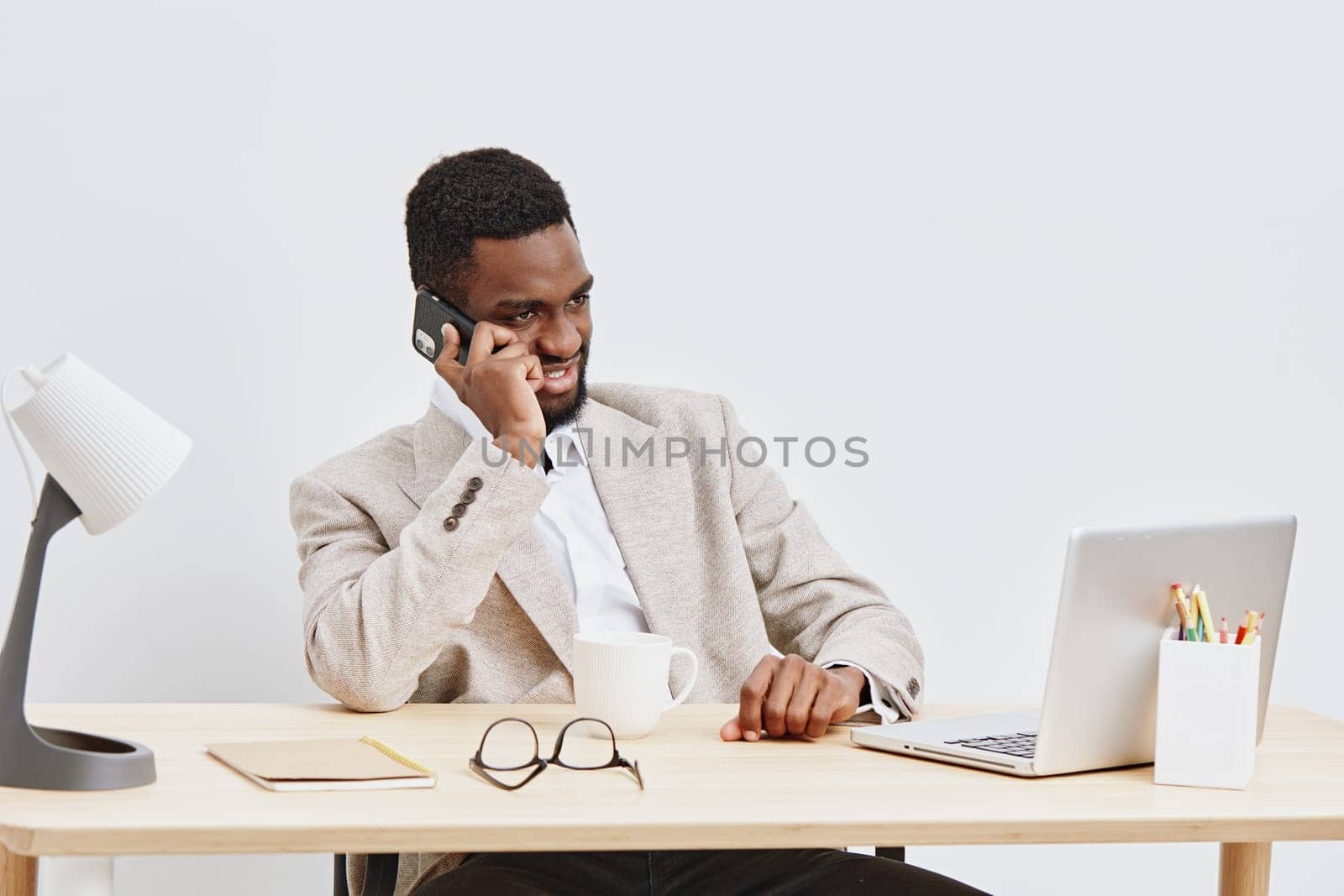 Concise image of a smiling young African American man in professional attire sitting at a desk with a laptop, holding a phone, surrounded by office supplies, expressing productivity and communication in a corporate setting. Concept: modern business communication