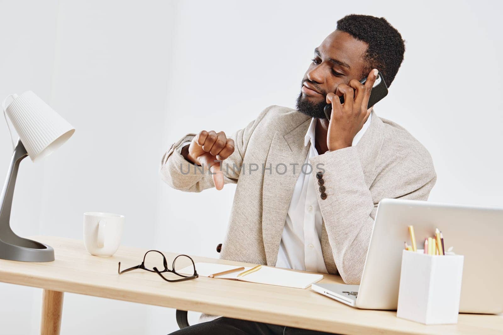 Confident man in smart casual attire using mobile phone, seated at an organized desk with laptop, glasses, and stationery. Bright office environment, focused expression, productivity, modern professional atmosphere.