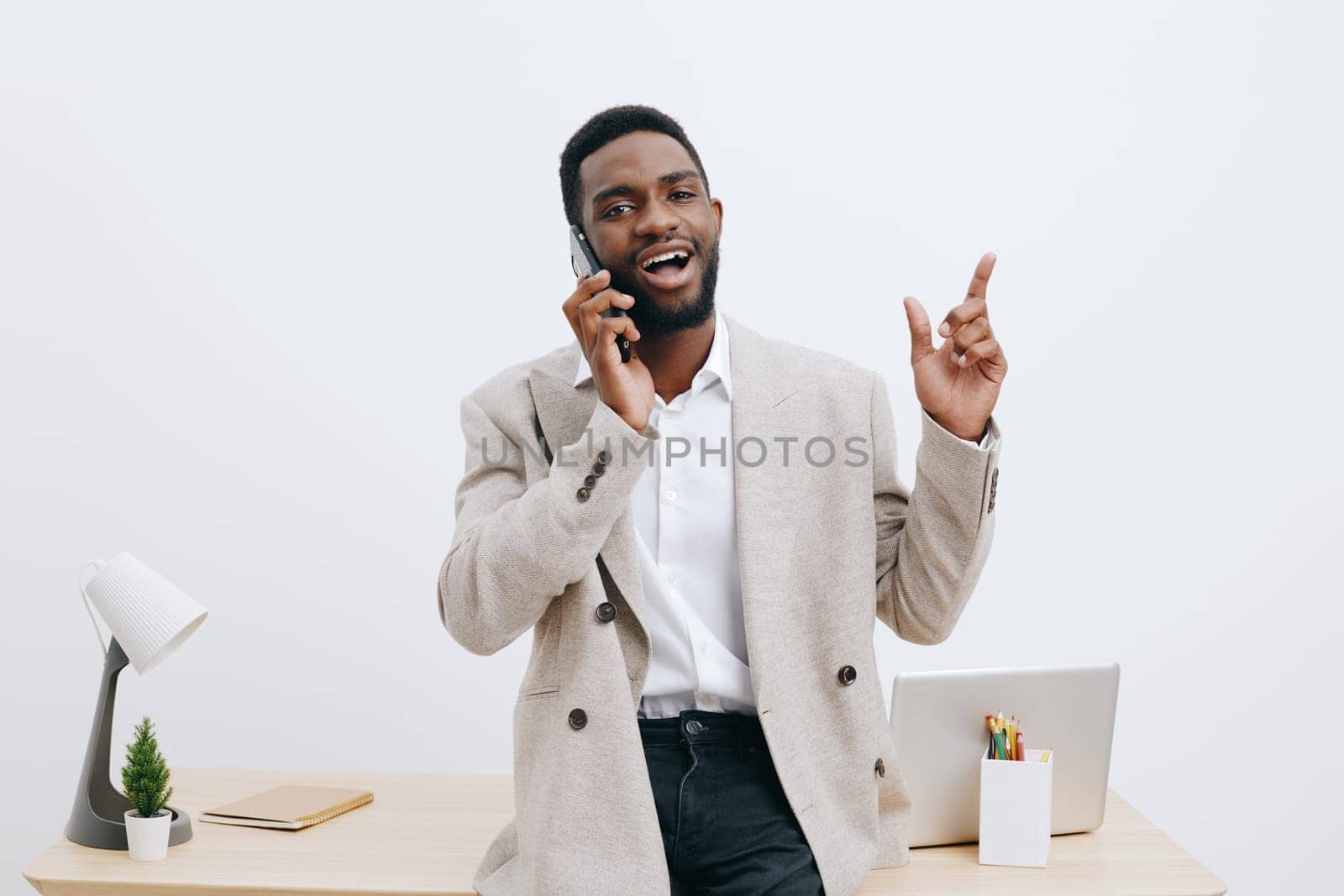 Confident smiling man engaging in a phone conversation while standing at a desk in a contemporary office environment with minimal decor, laptop, and stationery, illustrating effective communication, modern office, casual professional style, cheerful atmosphere, studio shot, concept: business communication.