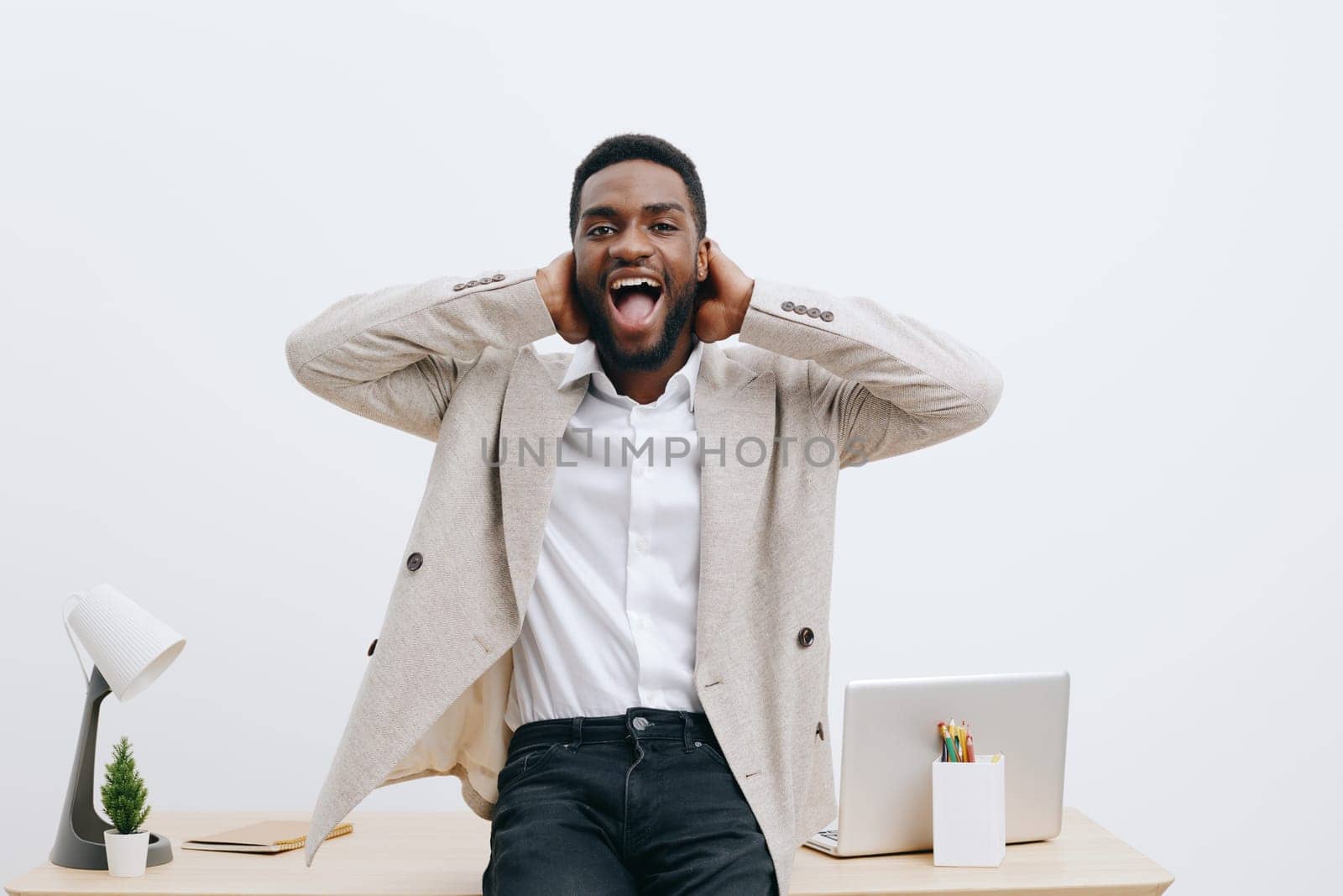 Joyful enthusiastic young man with dark skin and short hair, dressed in a beige blazer and white shirt, expressing excitement in bright contemporary office setting. Hands near ears, cheerful and energetic, workspace environment, happiness concept, lifestyle shot