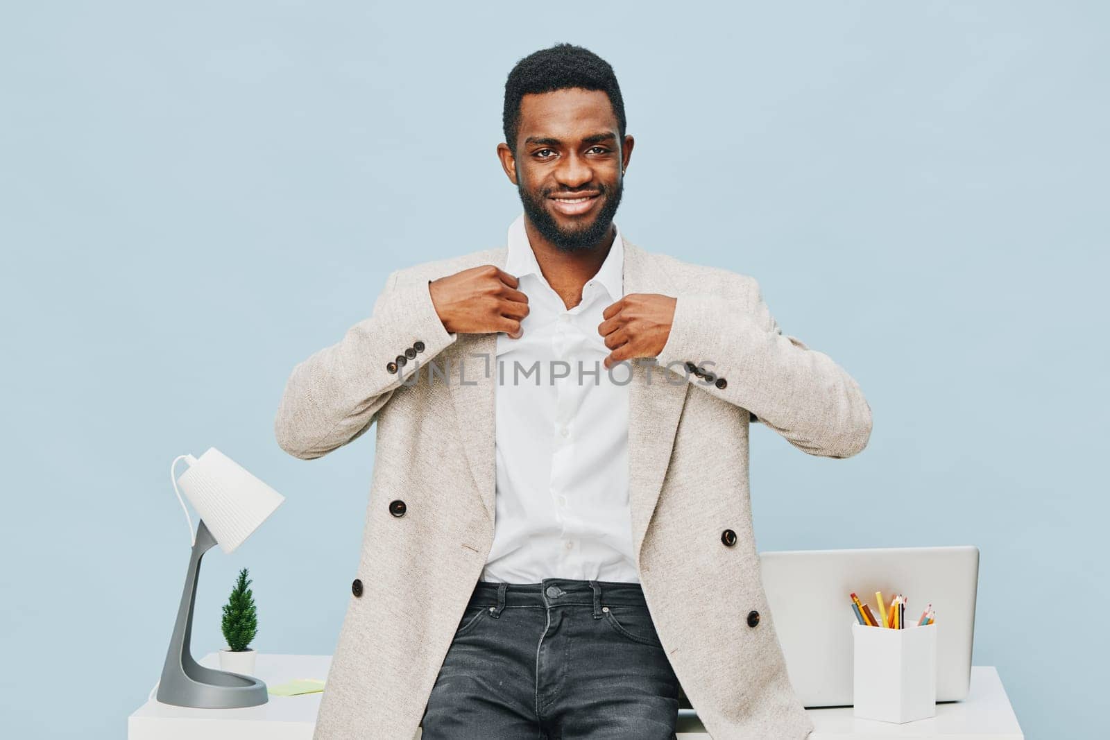 Confident smiling man in stylish beige blazer and white shirt standing at desk in contemporary office space with ergonomic chair, desk accessories, and sensor lighting. Bright, professional atmosphere, young professional lifestyle.